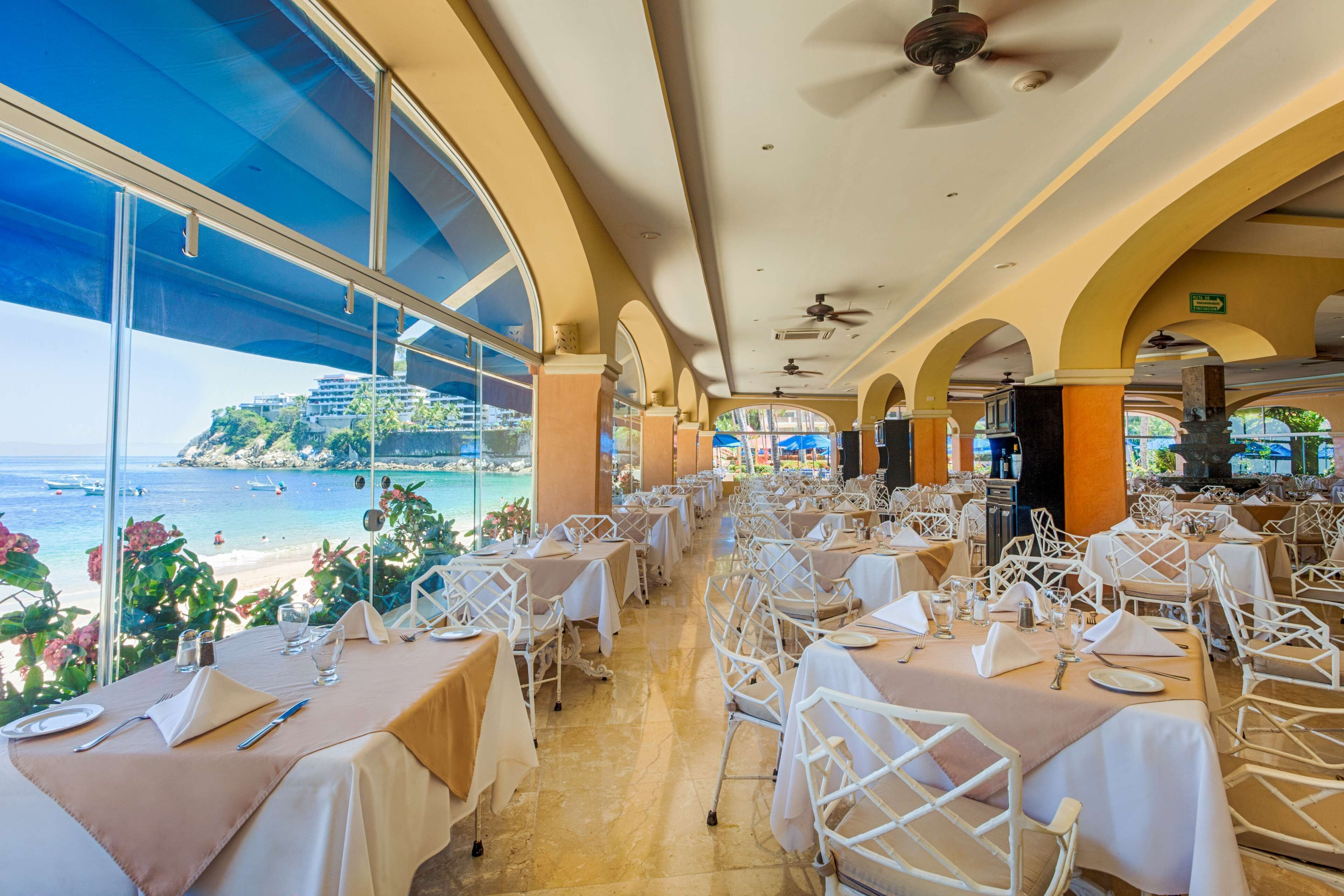A restaurant with tables set for dining, large windows offering a view of the beach and ocean, and a ceiling fan.