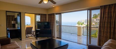 A spacious living room with a flat-screen TV, comfortable seating, a ceiling fan, and a balcony with a view of the ocean.