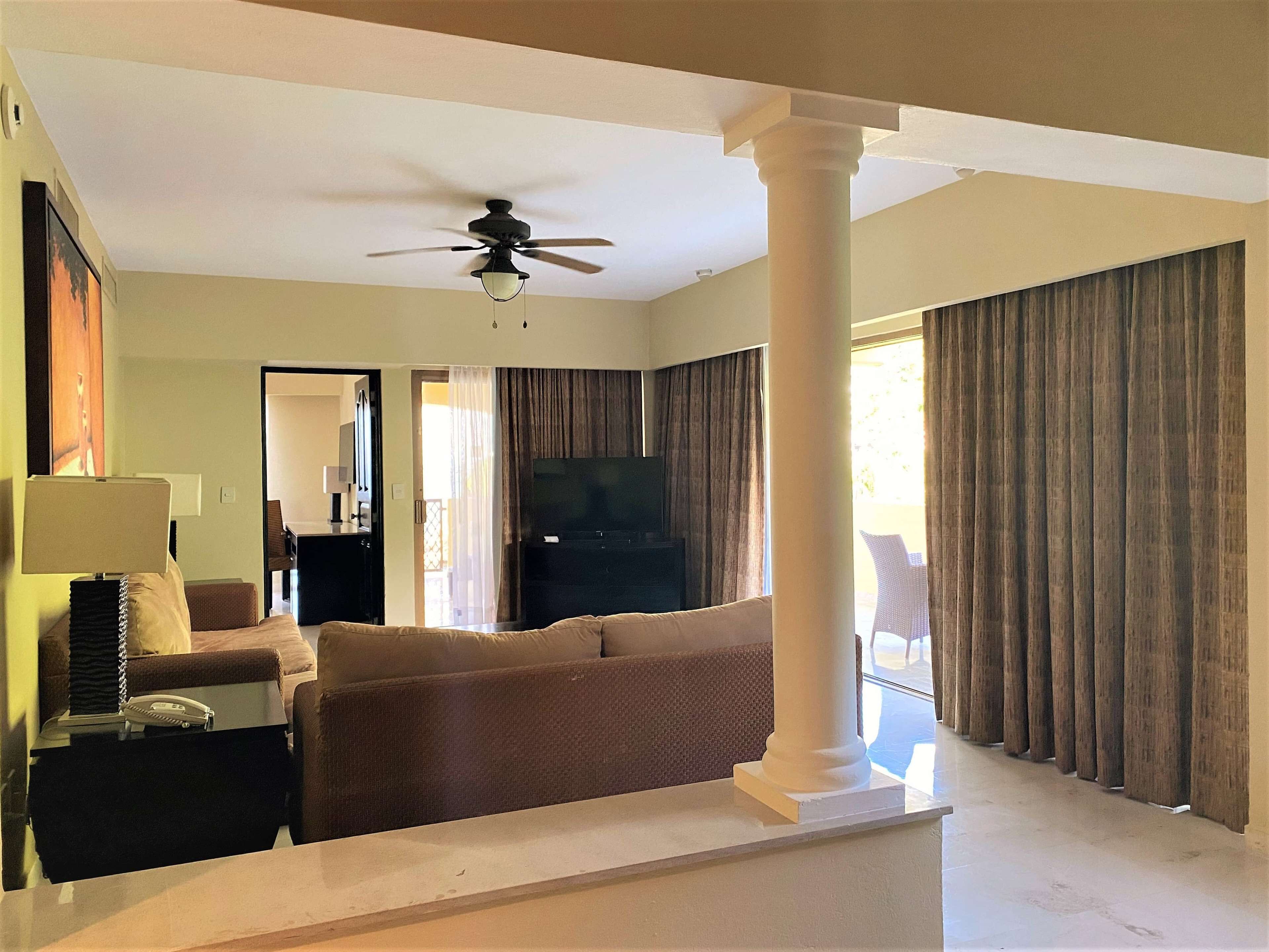 A spacious living room with a ceiling fan, a flat-screen TV, and a large window with curtains.