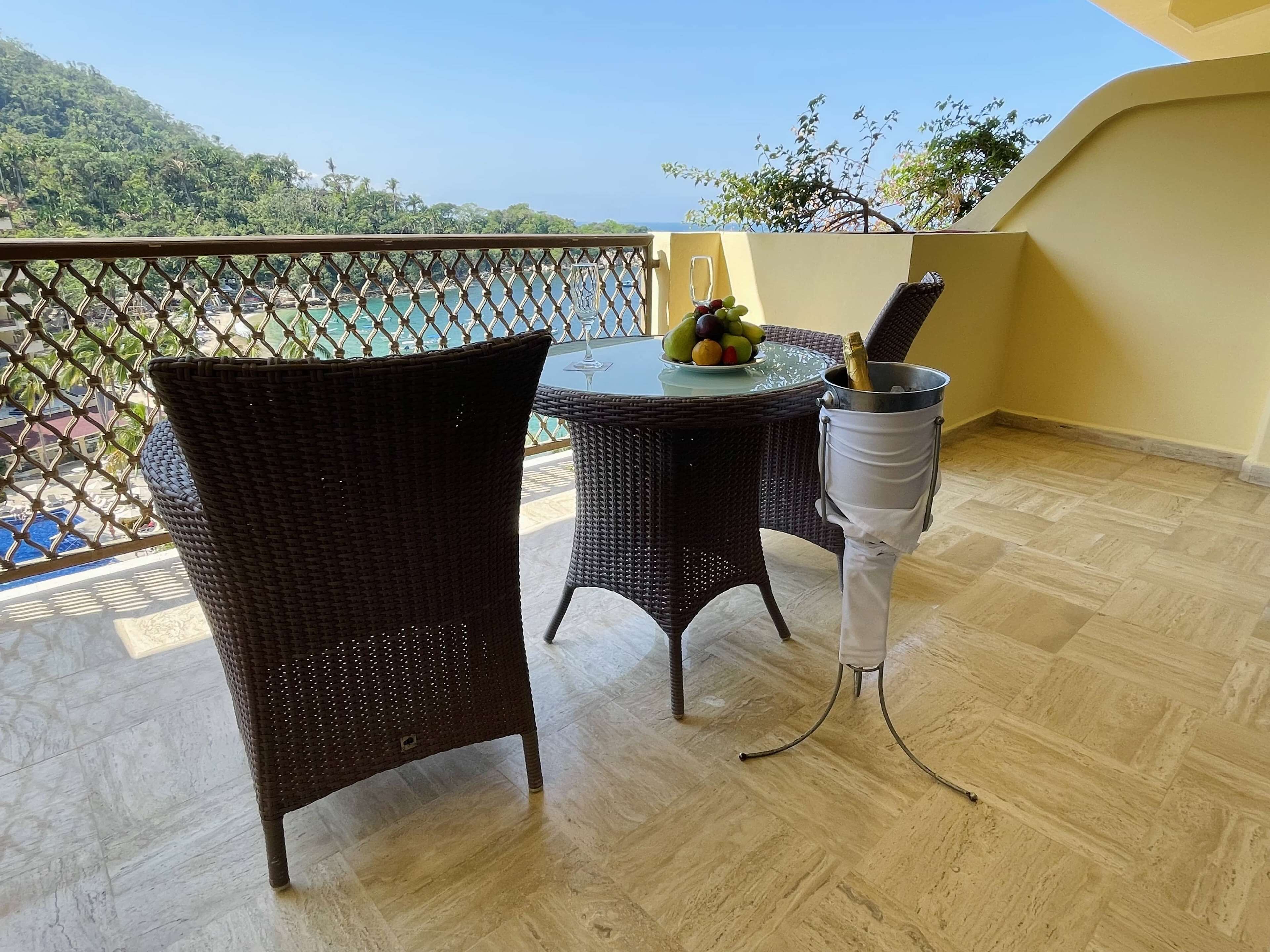 Un balcón con sillas de mimbre, una mesa con fruta y un cubo para champán, y vistas a una playa y montañas.