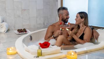 Una pareja en una bañera con pétalos de rosa y velas, rodeada de toallas y un plato de uvas.