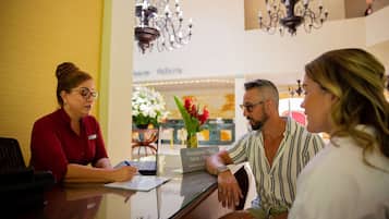 Un comptoir d’accueil avec une pancarte indiquant les services proposés, tels que des visites guidées et des réservations. Des lustres sont suspendus au plafond et un vase rempli de fleurs est posé sur le comptoir.