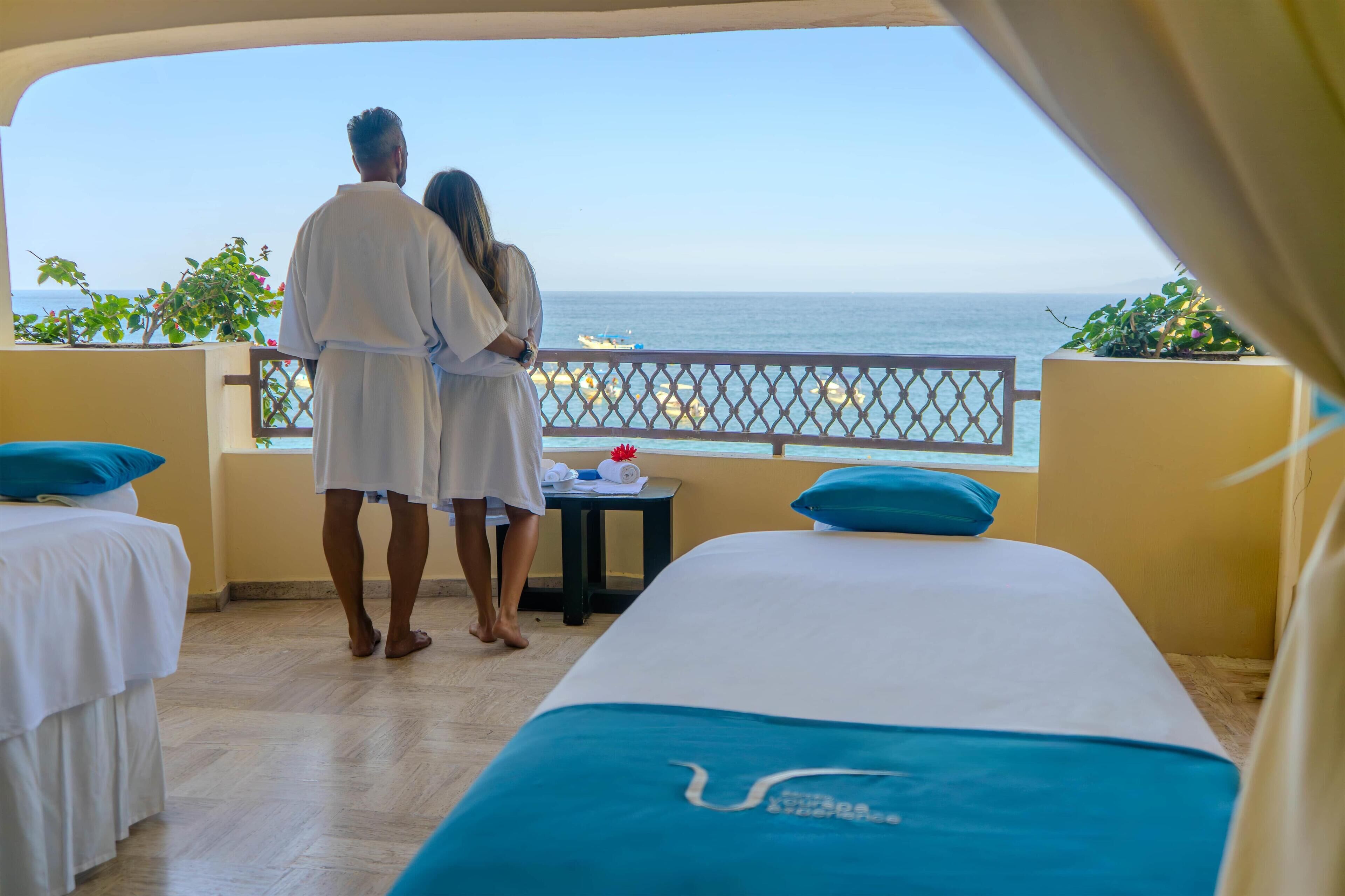 Ein Paar steht an einem Balkon mit Blick aufs Meer, mit einem Bett und einem kleinen Tisch mit einem Handtuch und einer roten Blume.