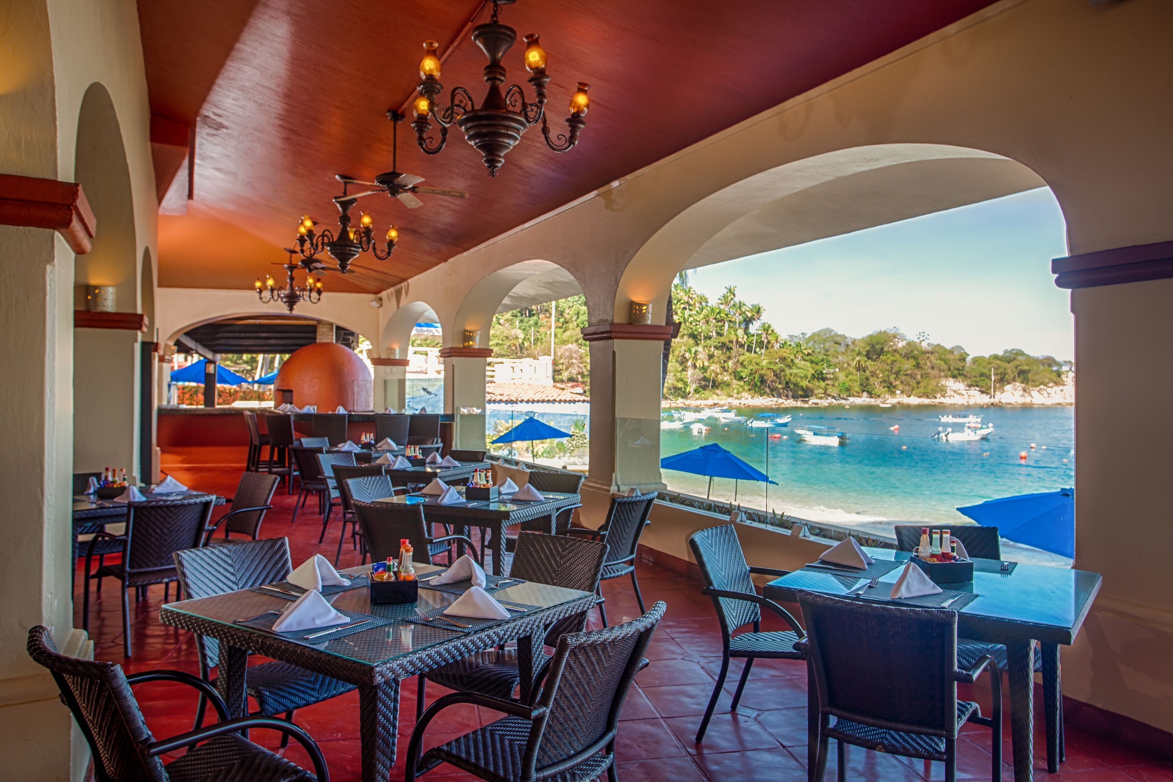 Un área de comedor al aire libre con mesas y sillas, vista al mar y sombrillas.