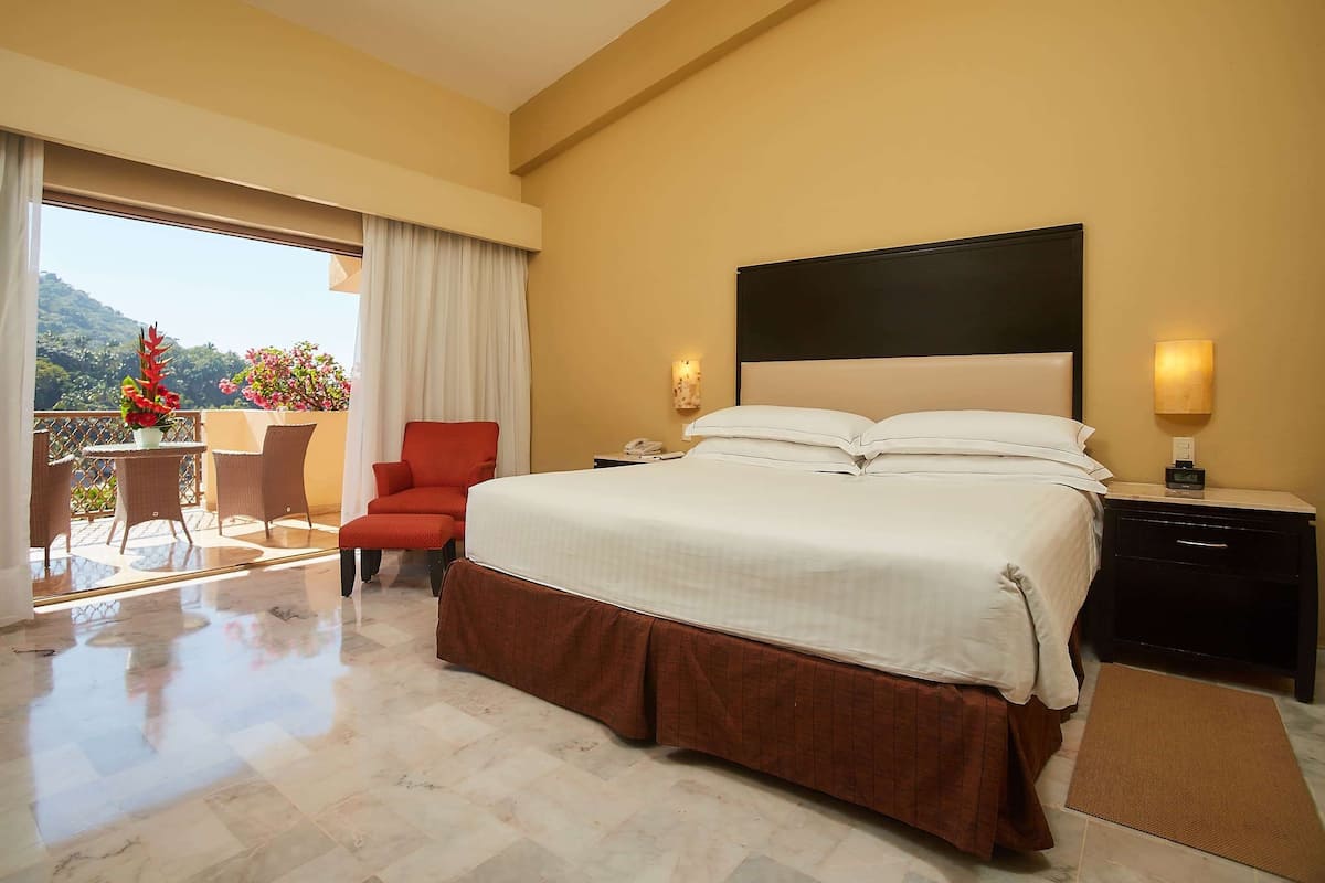 A hotel room with a large bed, a nightstand, a chair, a balcony with a table and chairs, and a view of the outdoors.