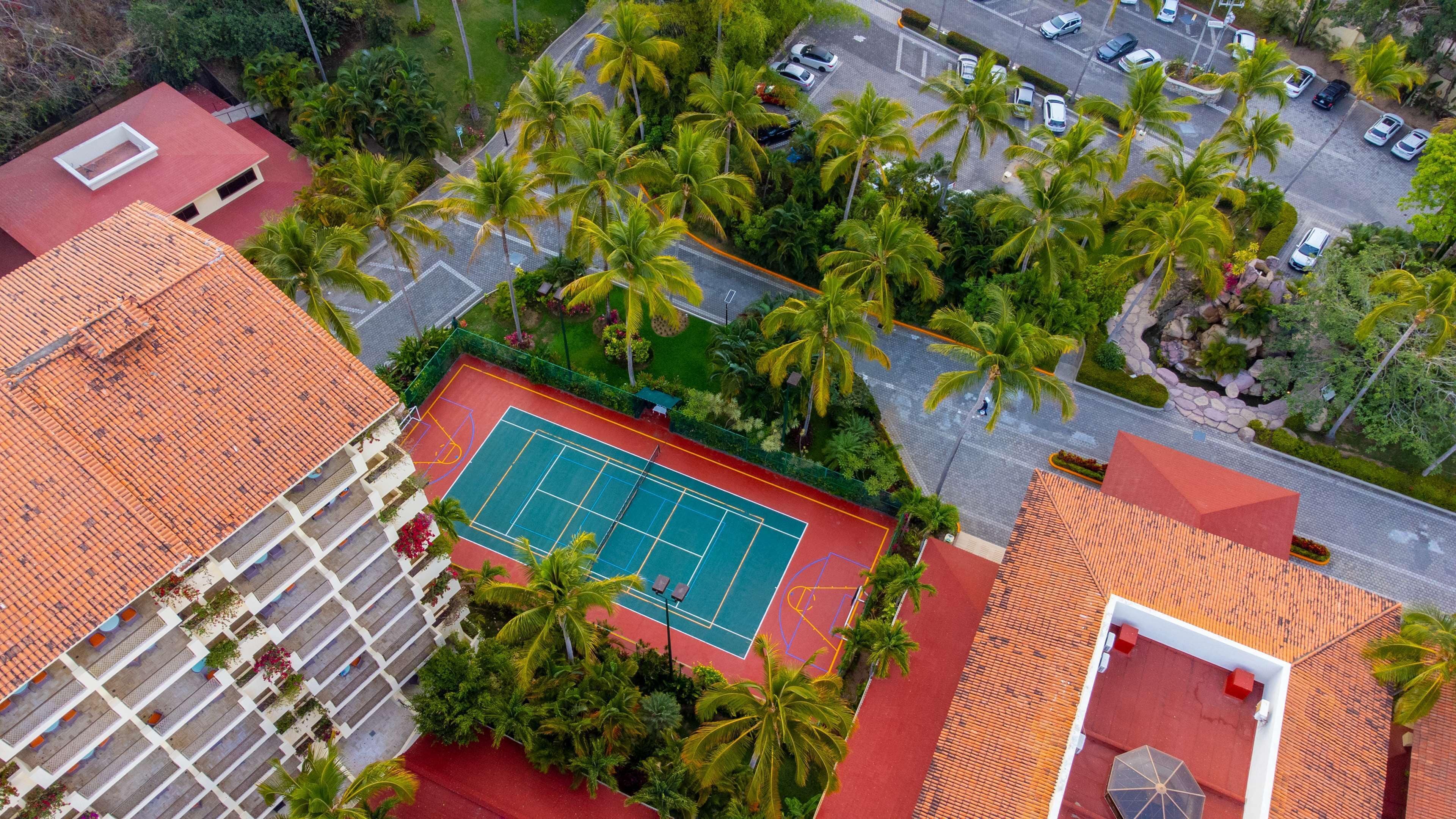 An aerial view of a residential area with a tennis court surrounded by palm trees and a parking lot.