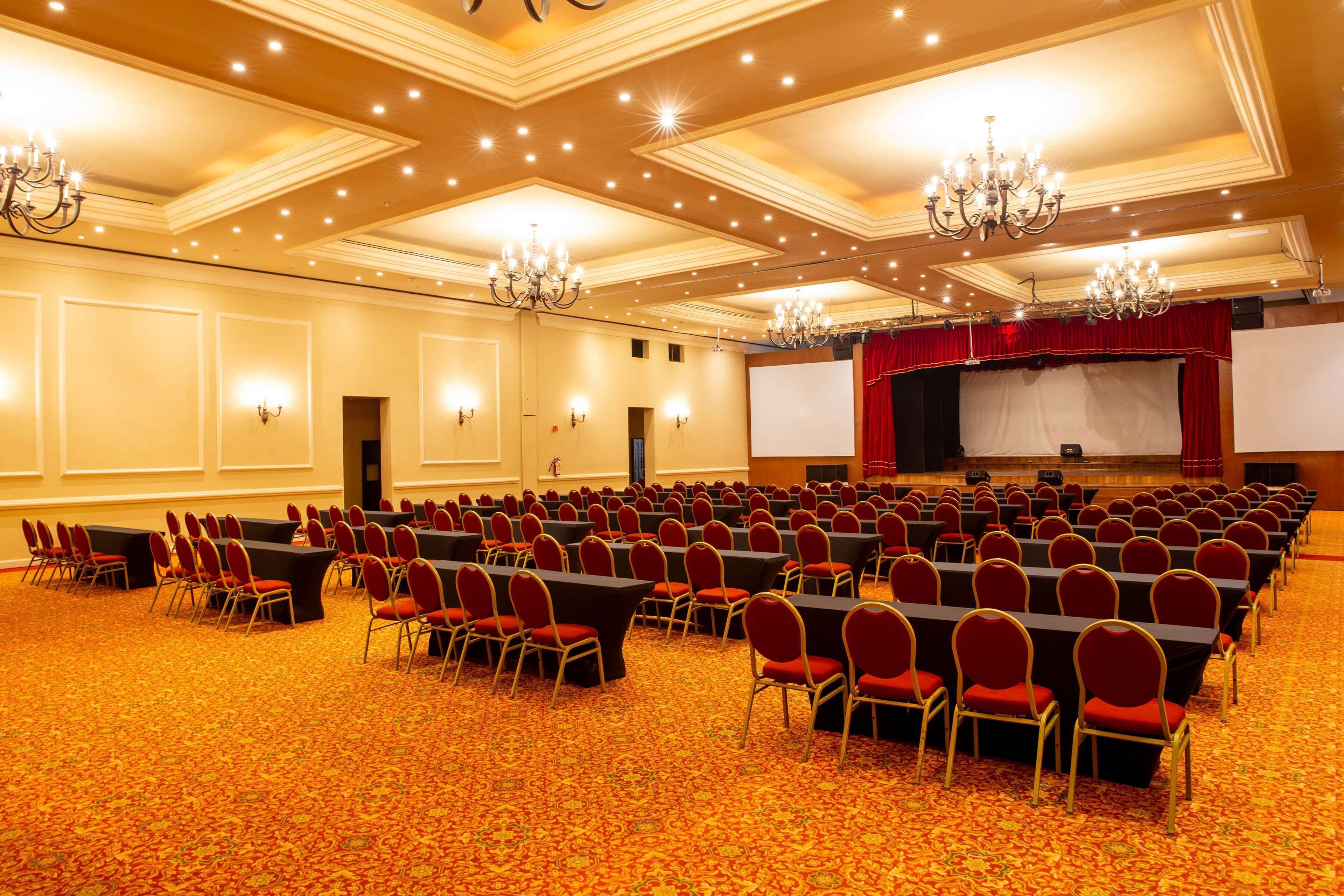 Un salón de conferencias amplio con filas de sillas, un escenario con cortina roja y candelabros.