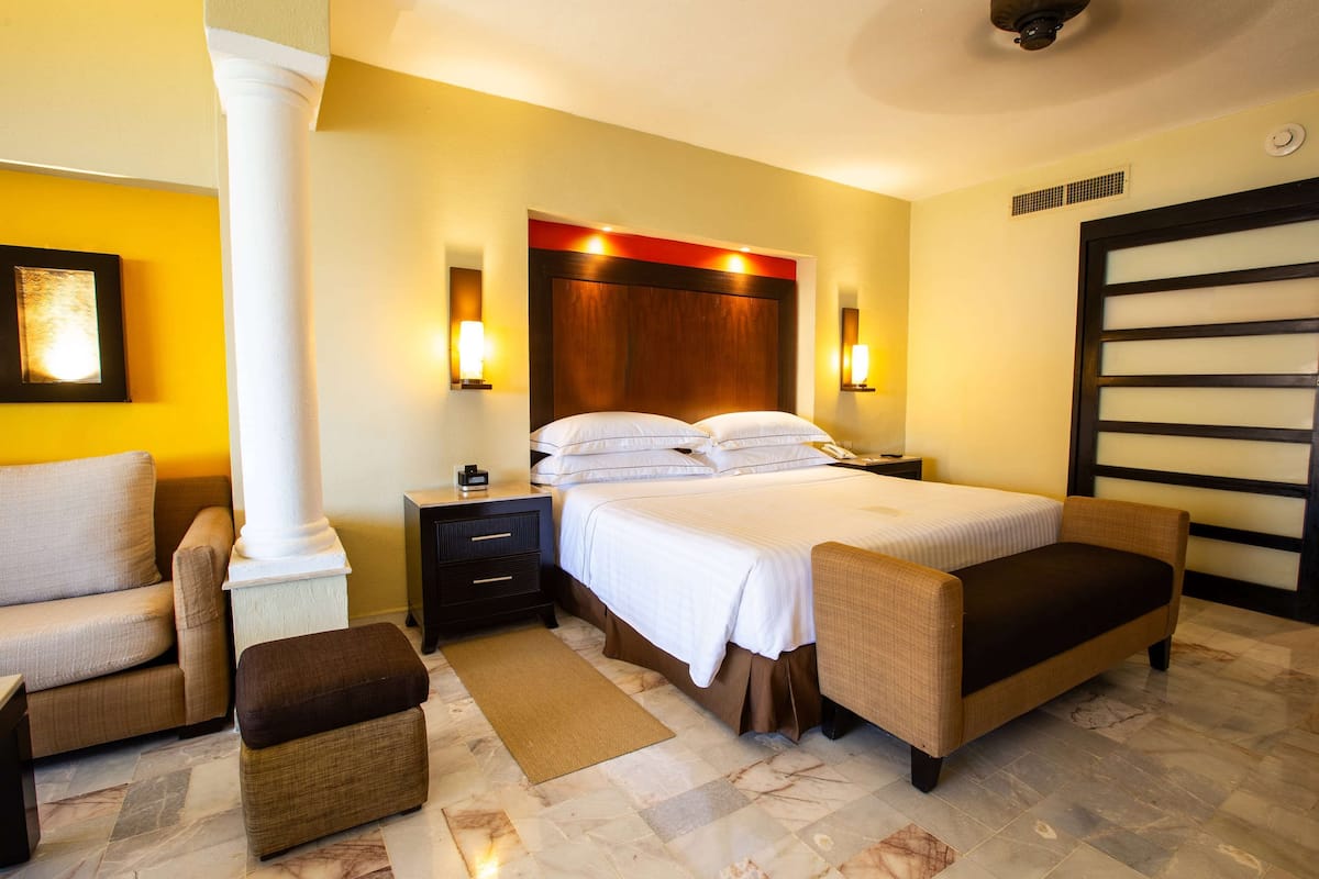 A hotel room with a large bed, two bedside tables, a nightstand, a sofa, an ottoman, and a large window with blinds.