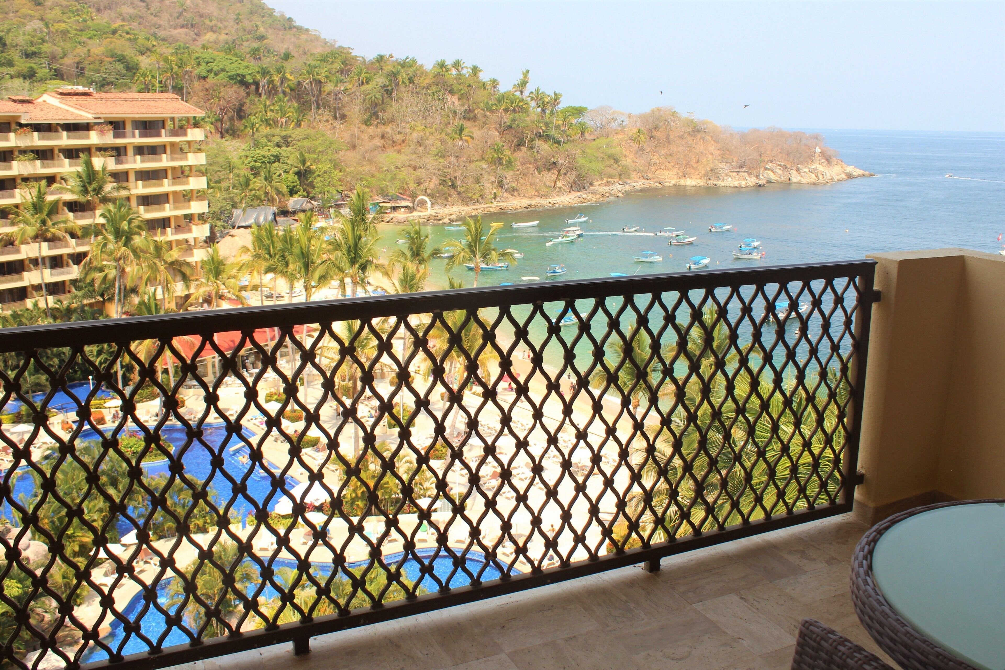 A balcony with a view of a beach, a pool, and a building in the background.