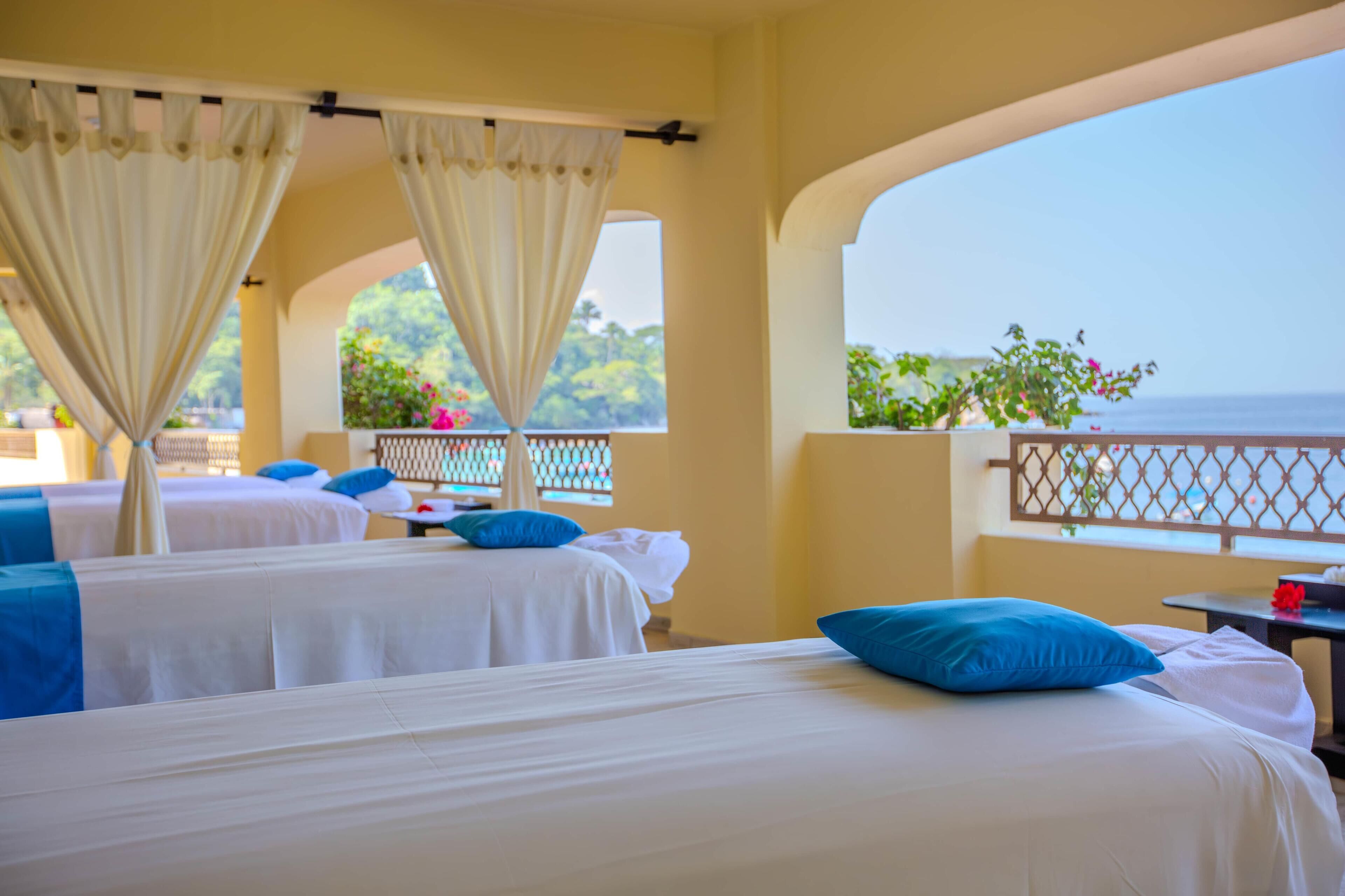 A hotel room with two beds, each with white linens and blue pillows, a balcony with a view of the sea, and a window with curtains.