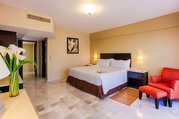 Una habitación de hotel con cama, mesitas de noche, una silla roja, un jarrón de flores y un cuadro en la pared.