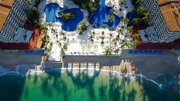 An aerial view of a resort with multiple swimming pools, palm trees, and beach access.