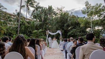 Una ceremonia de boda al aire libre con invitados sentados en sillas blancas, mirando a una pareja debajo de un arco blanco.