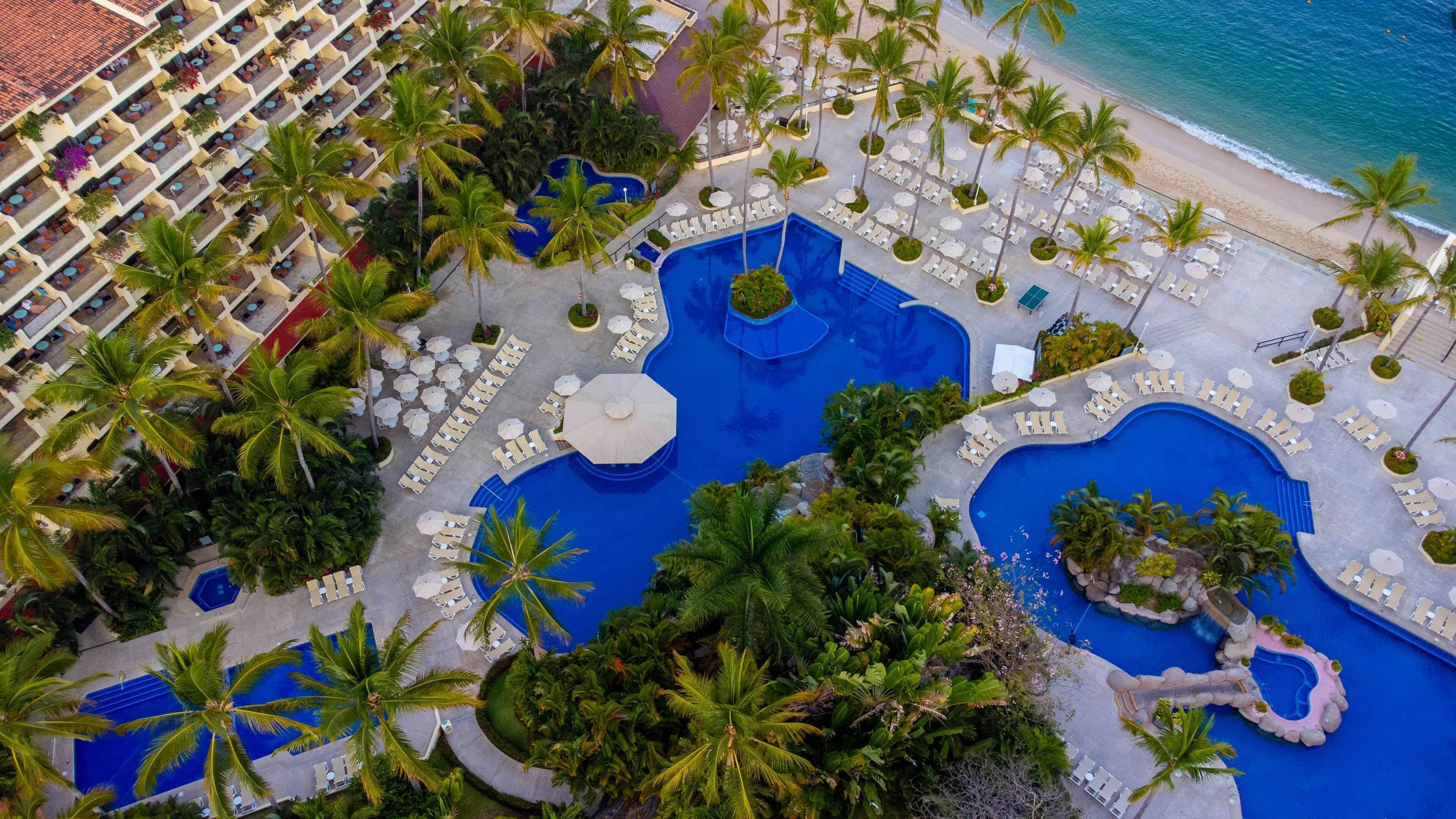 An aerial view of a resort with multiple swimming pools, palm trees, and beach access.