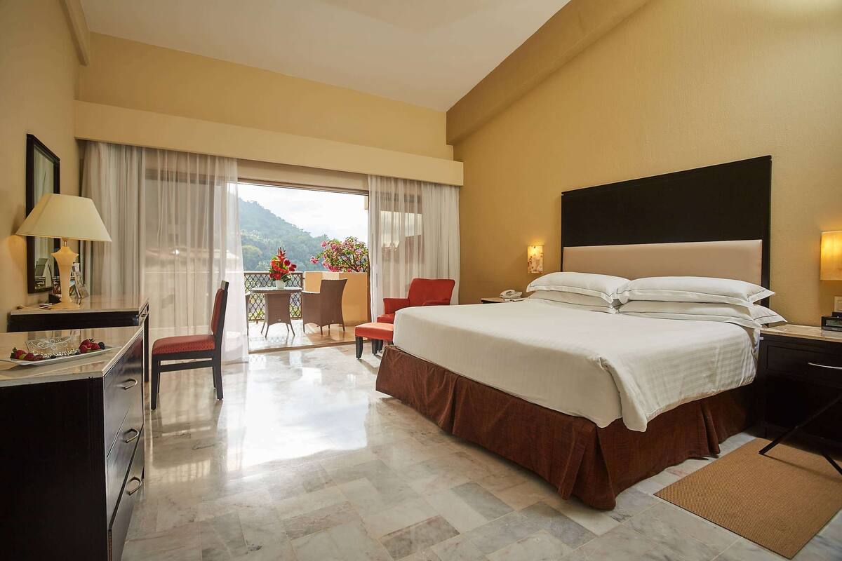 A hotel room with a bed, a desk with a lamp, a chair, a small table with flowers, and a view of the outdoors.
