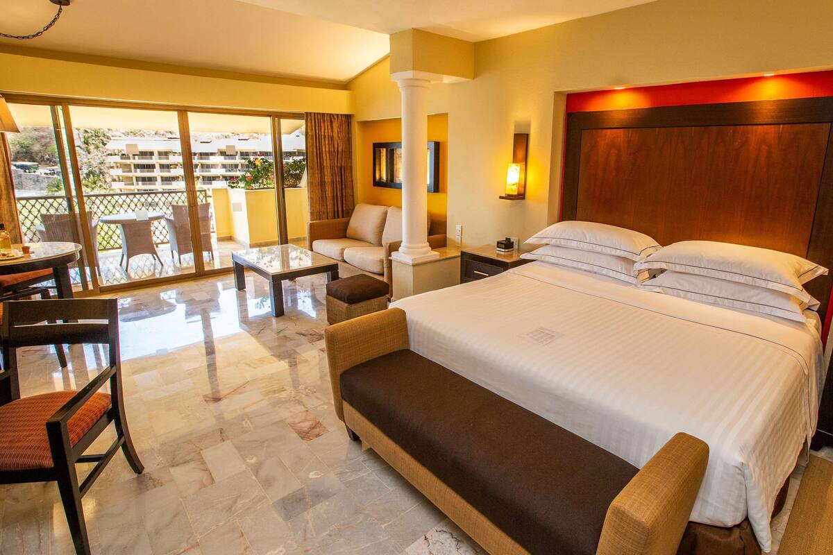 A hotel room with a large bed, a sitting area with a sofa and chairs, a small table, and a balcony with a view of the surrounding area.