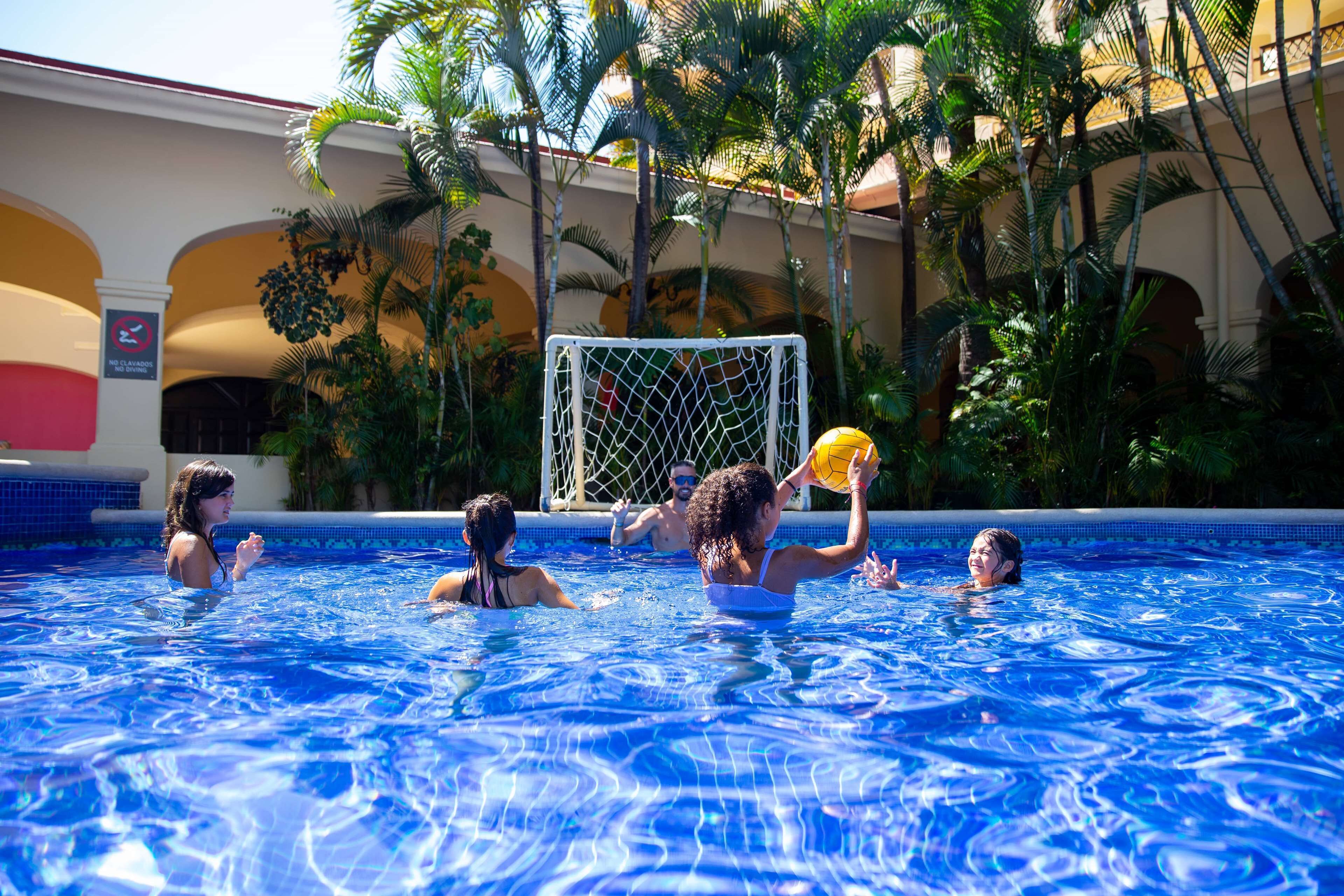 A swimming pool with people playing with a ball, surrounded by palm trees and a building with arches.