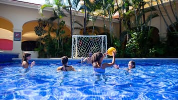 A swimming pool with people playing with a ball, surrounded by palm trees and a building with arches.