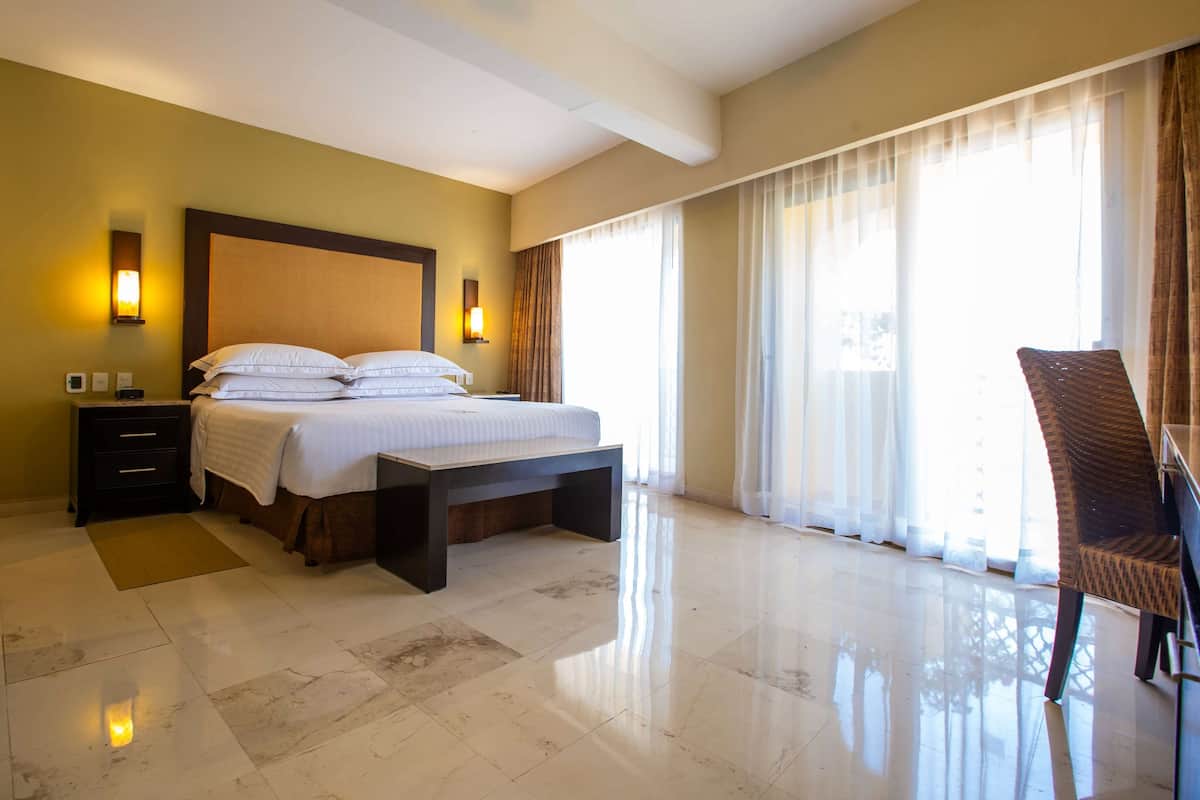 A hotel room with a bed, bedside tables, a bench, a chair, and large windows with curtains.