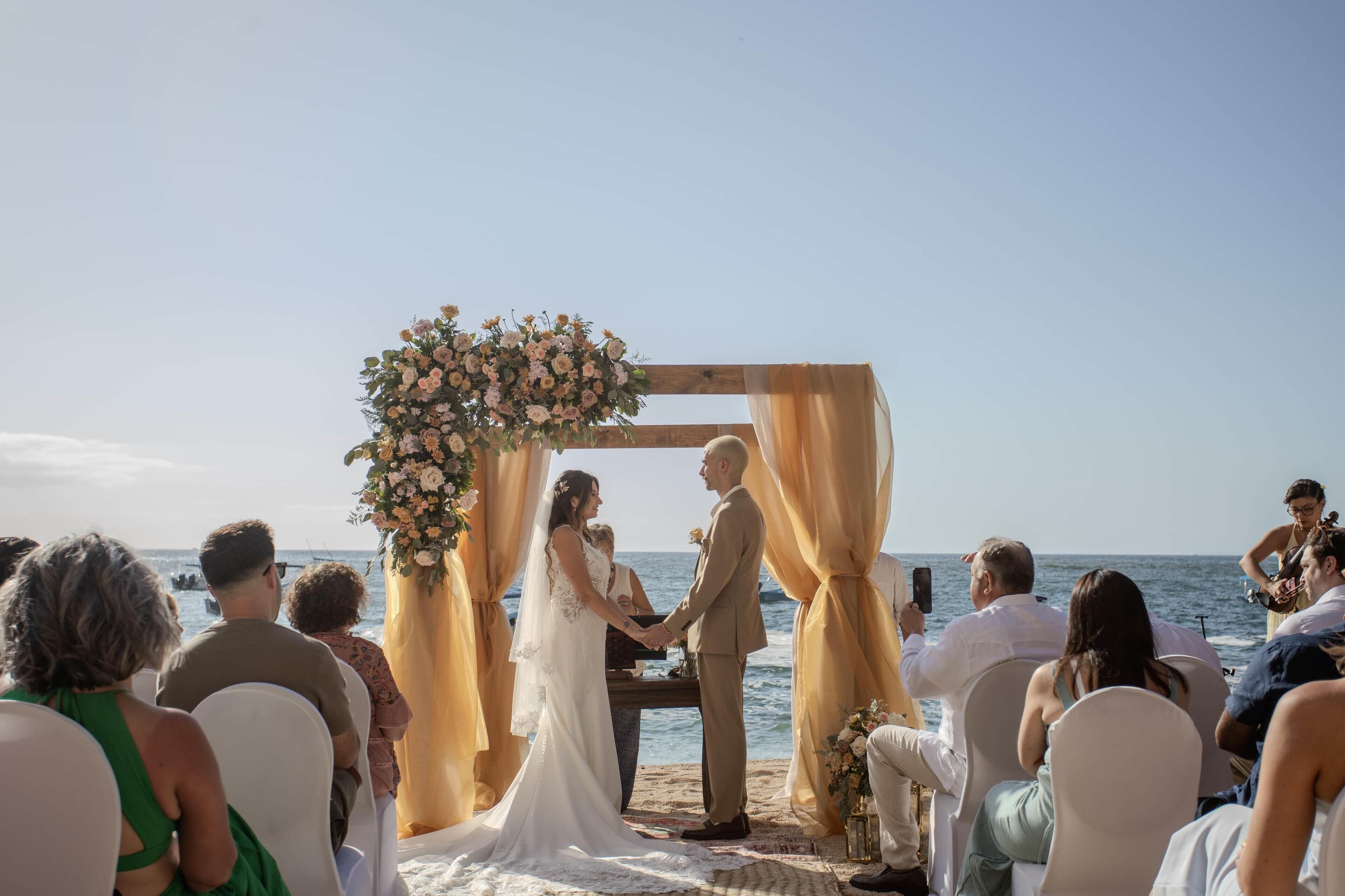 A wedding ceremony on a beach with a wooden arch decorated with flowers, guests seated on white chairs, and a clear view of the ocean.