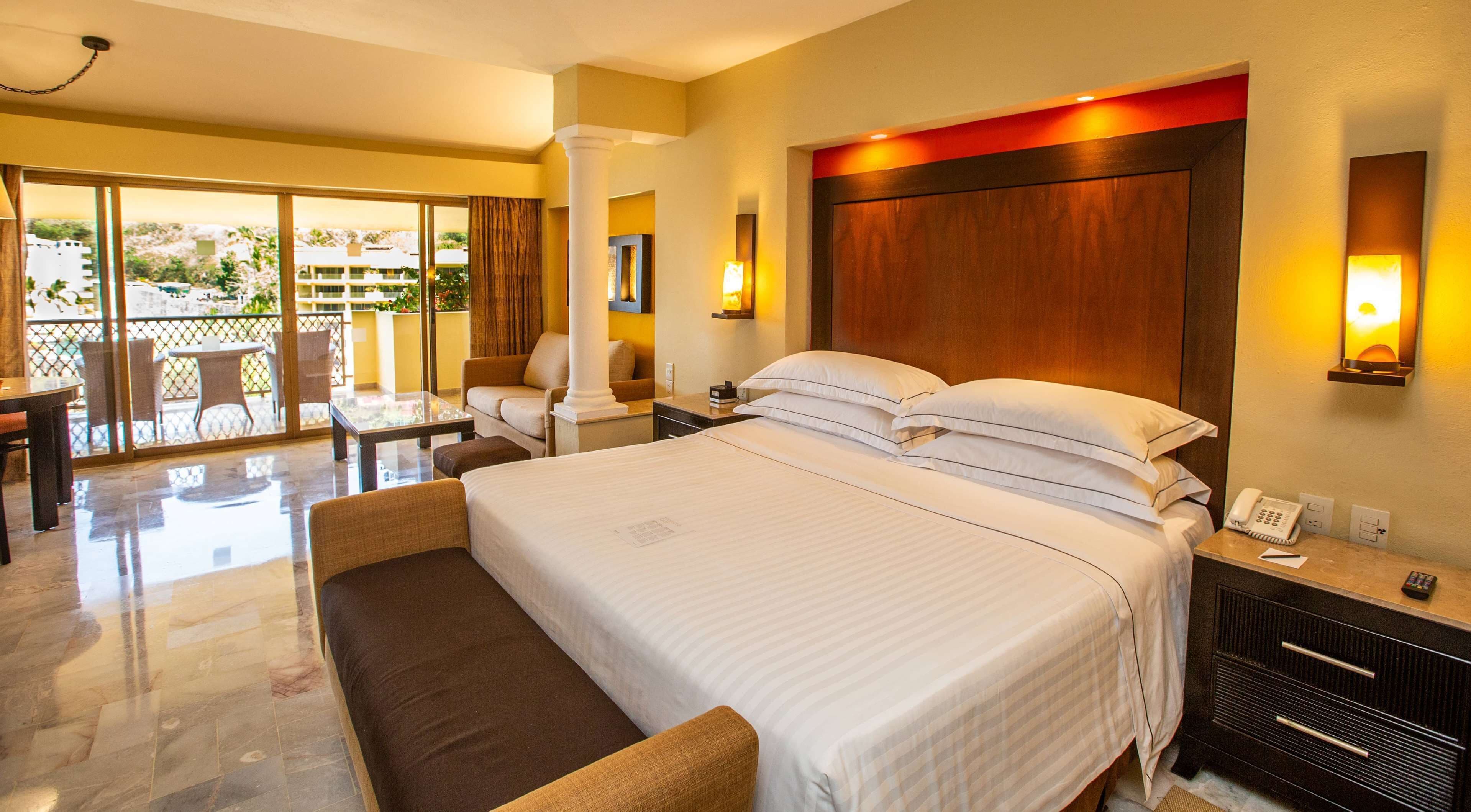 Habitación de hotel con una cama grande, un área de descanso con sofá y sillones, una mesita y un balcón con vistas al exterior.