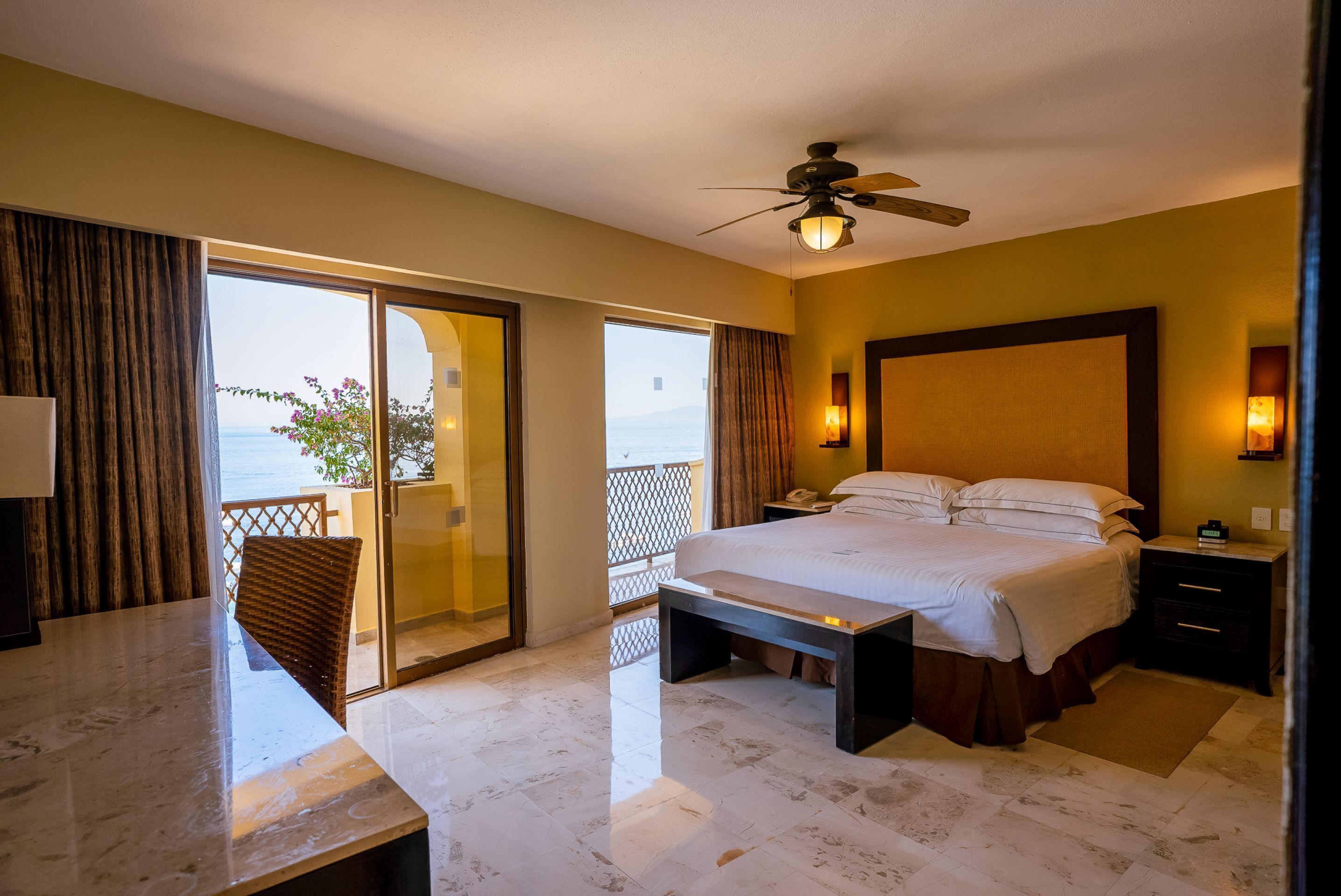 A hotel room with a bed, a desk, and a balcony with a view of the sea.