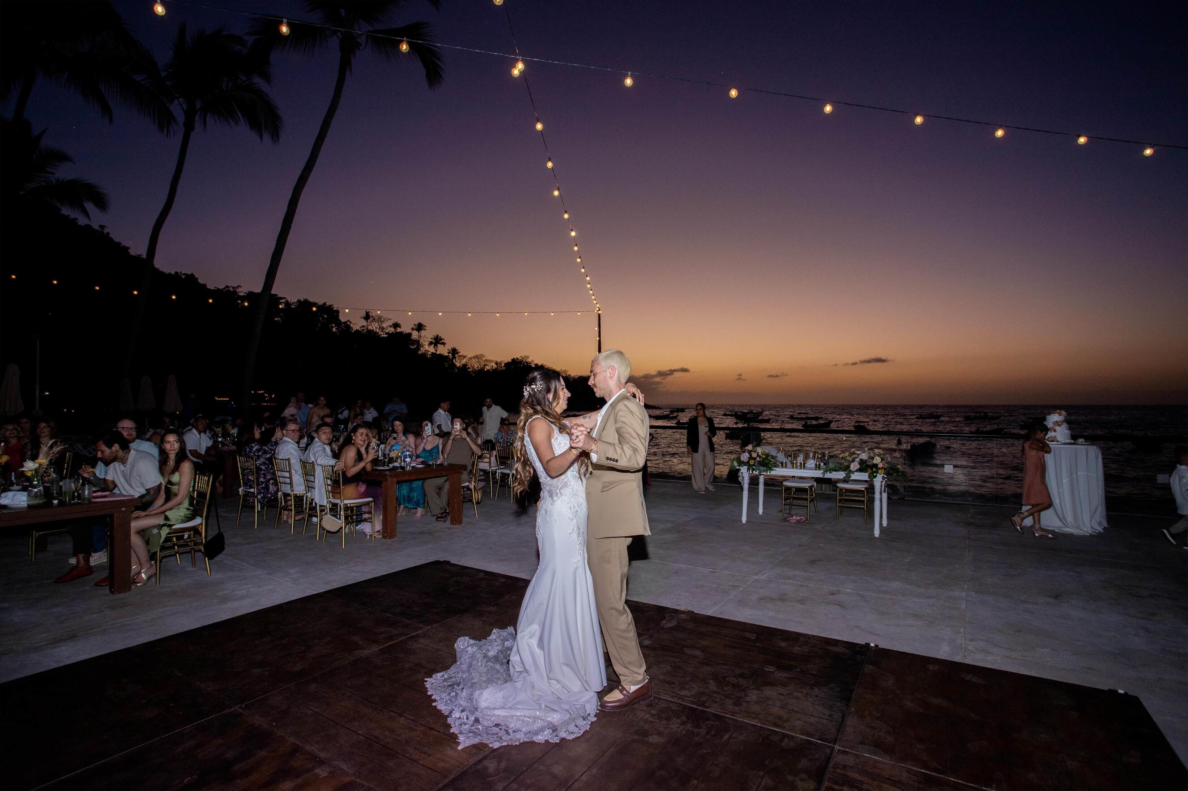 A wedding ceremony with a couple dancing on a wooden dance floor, guests seated at tables, and a view of the ocean.