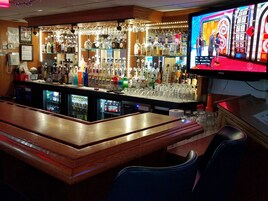 Zona de bar con mostrador, estanterías llenas de botellas de licor y una pantalla de televisión mostrando un partido.