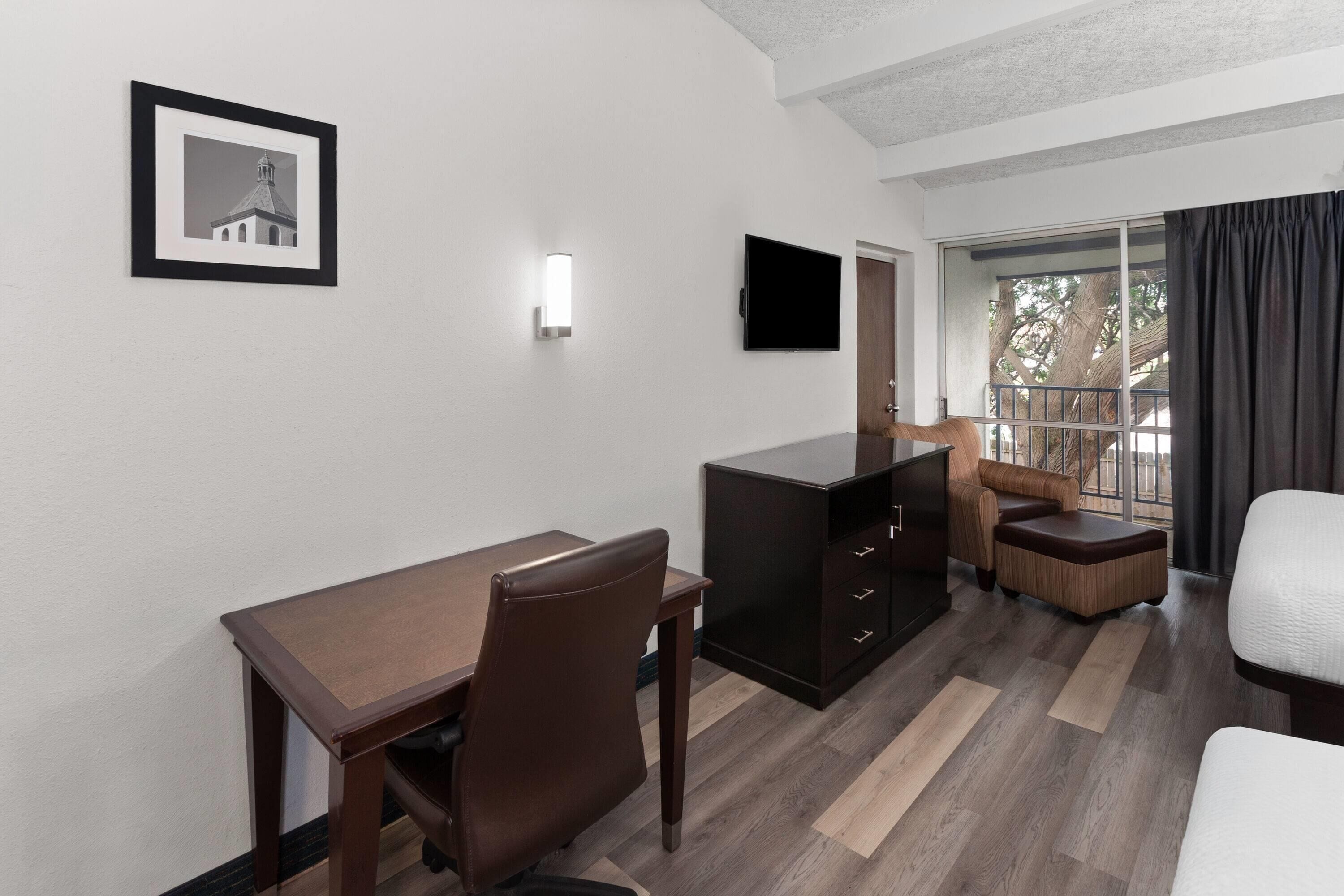 A hotel room with a desk, chair, sofa, and a television mounted on the wall.