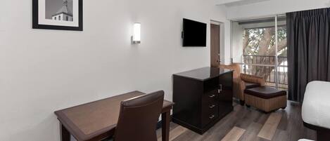 A hotel room with a desk, chair, sofa, and a television mounted on the wall.