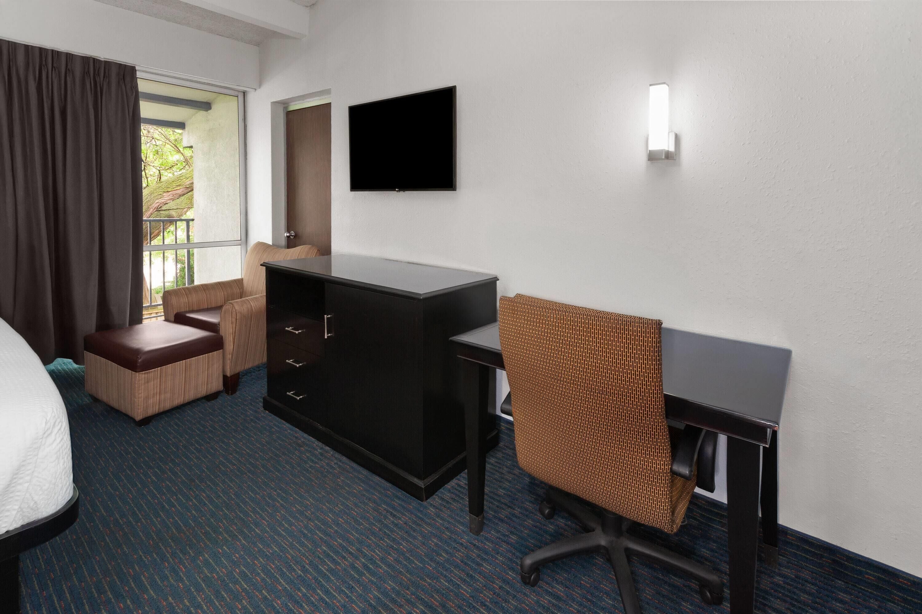 Habitación de hotel con cama, escritorio, silla, cómoda y televisor en la pared.