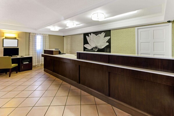 Un espace d’accueil comprenant un comptoir de réception en bois, une grande décoration florale murale, un téléviseur à écran plat et un coin salon avec une chaise et une petite table.