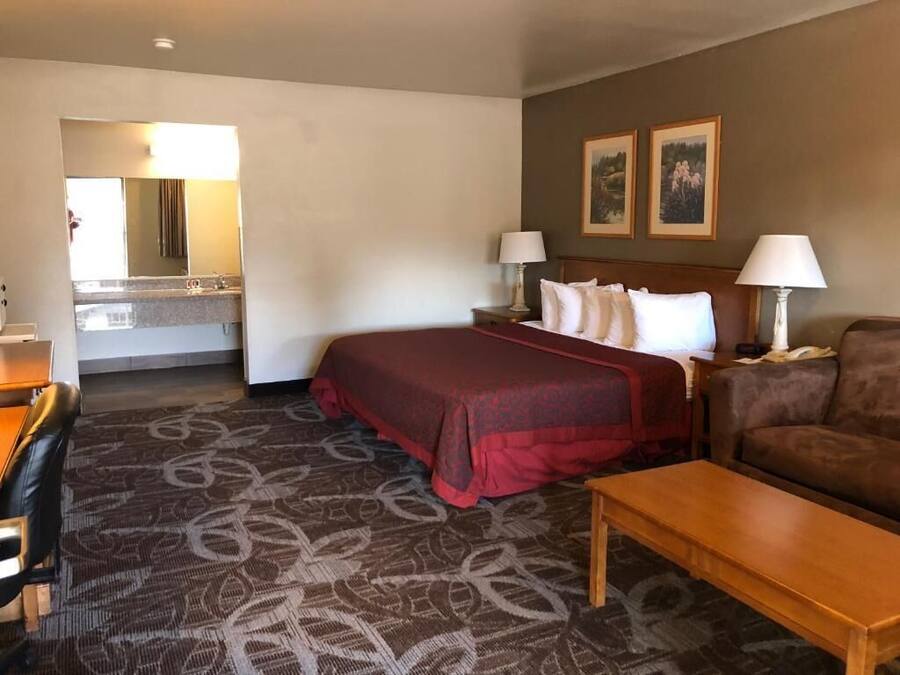 A hotel room with a bed, two bedside lamps, a wooden nightstand, a brown sofa, a wooden coffee table, a patterned carpet, a desk with a chair, and a bathroom with a mirror and sink.