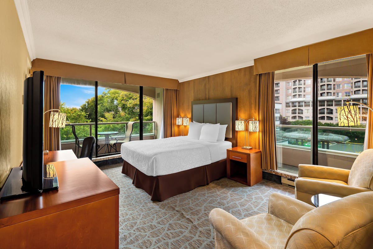 A hotel room with a large bed, a desk, a chair, and a balcony with a view of buildings and greenery.