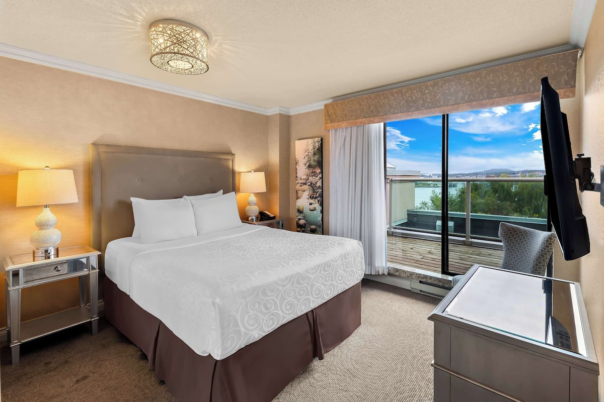 A hotel room with a large bed, two bedside lamps, a nightstand, a dresser, and a view of the outdoors through a large window.