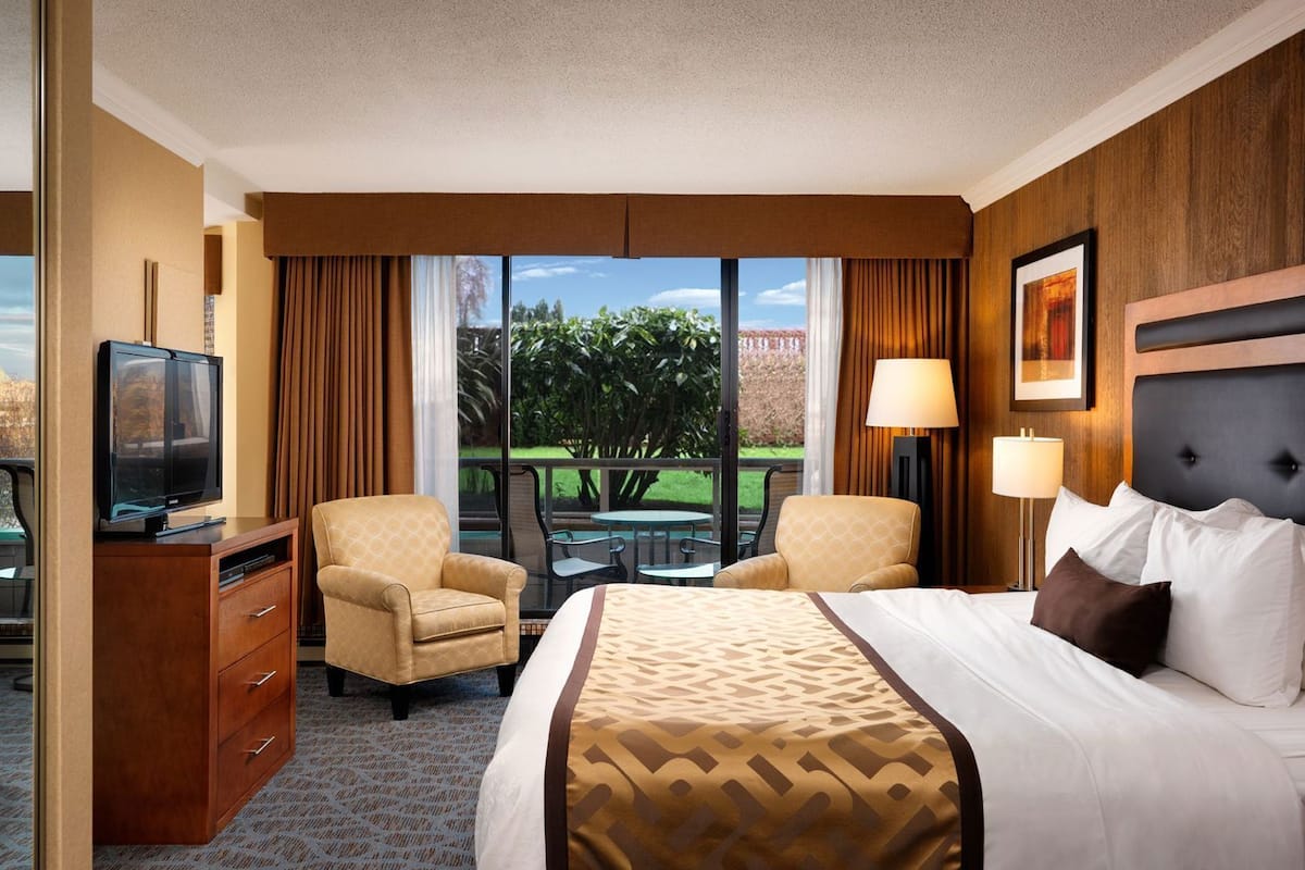 A hotel room with a bed, two armchairs, a TV, a desk, and a view of the outdoors.