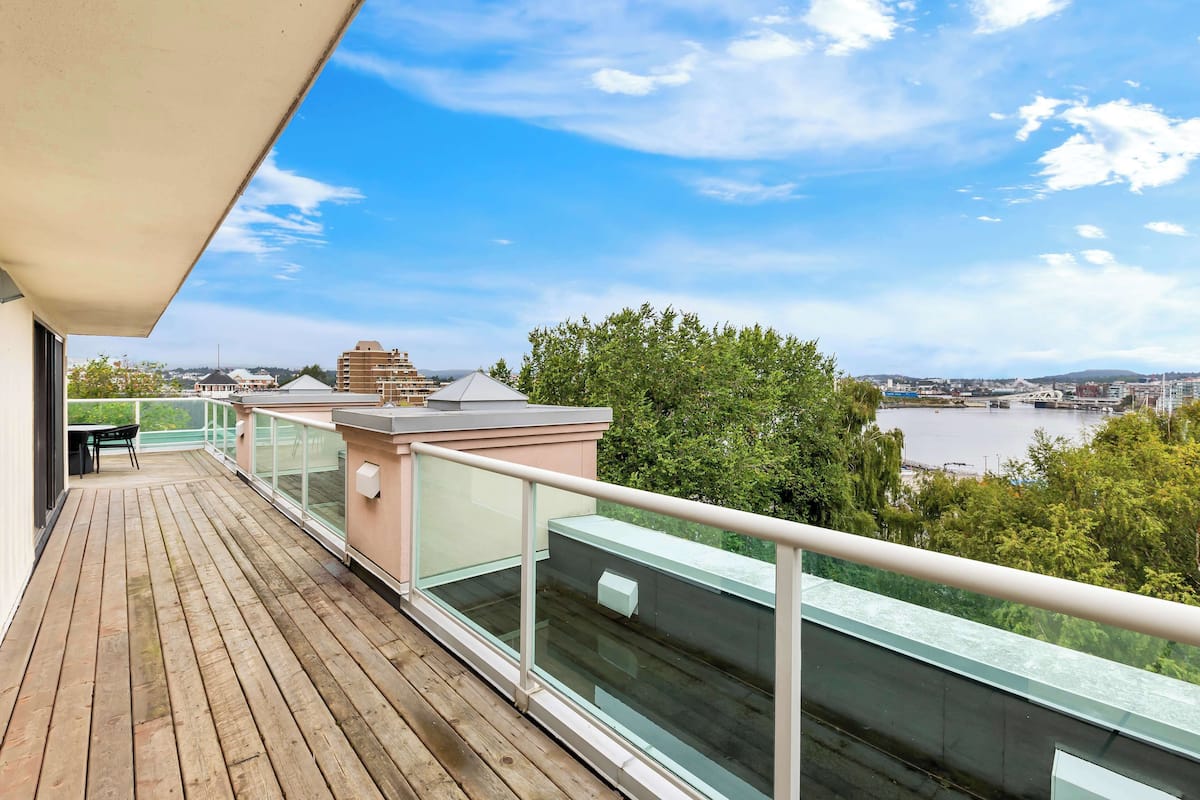 A wooden deck with a glass balustrade overlooking a river and cityscape.