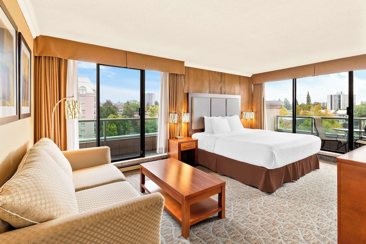 A hotel room with a bed, sofa, nightstand, lamp, and a view of the city.