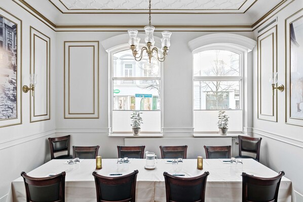 A dining room with a long table, six chairs, a chandelier, and two windows with potted plants.