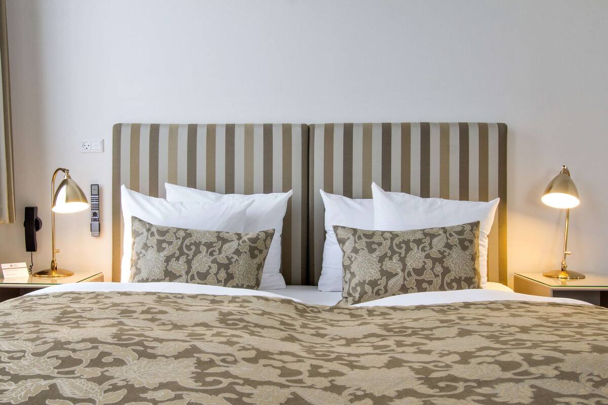 Una habitación de hotel con cabecera a rayas, dos lámparas de noche, una colcha estampada y una ventana con cortinas.
