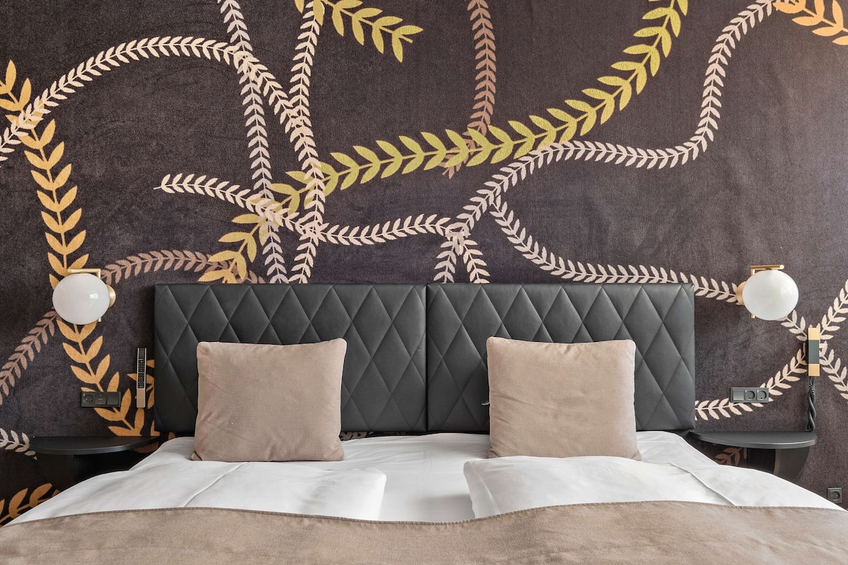 Una cama con cabecera capitonada, dos mesitas de noche con lámparas y una pared con estampado de hojas.