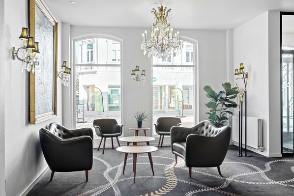A modern interior with leather armchairs, a chandelier, and large windows offering a view of the street.