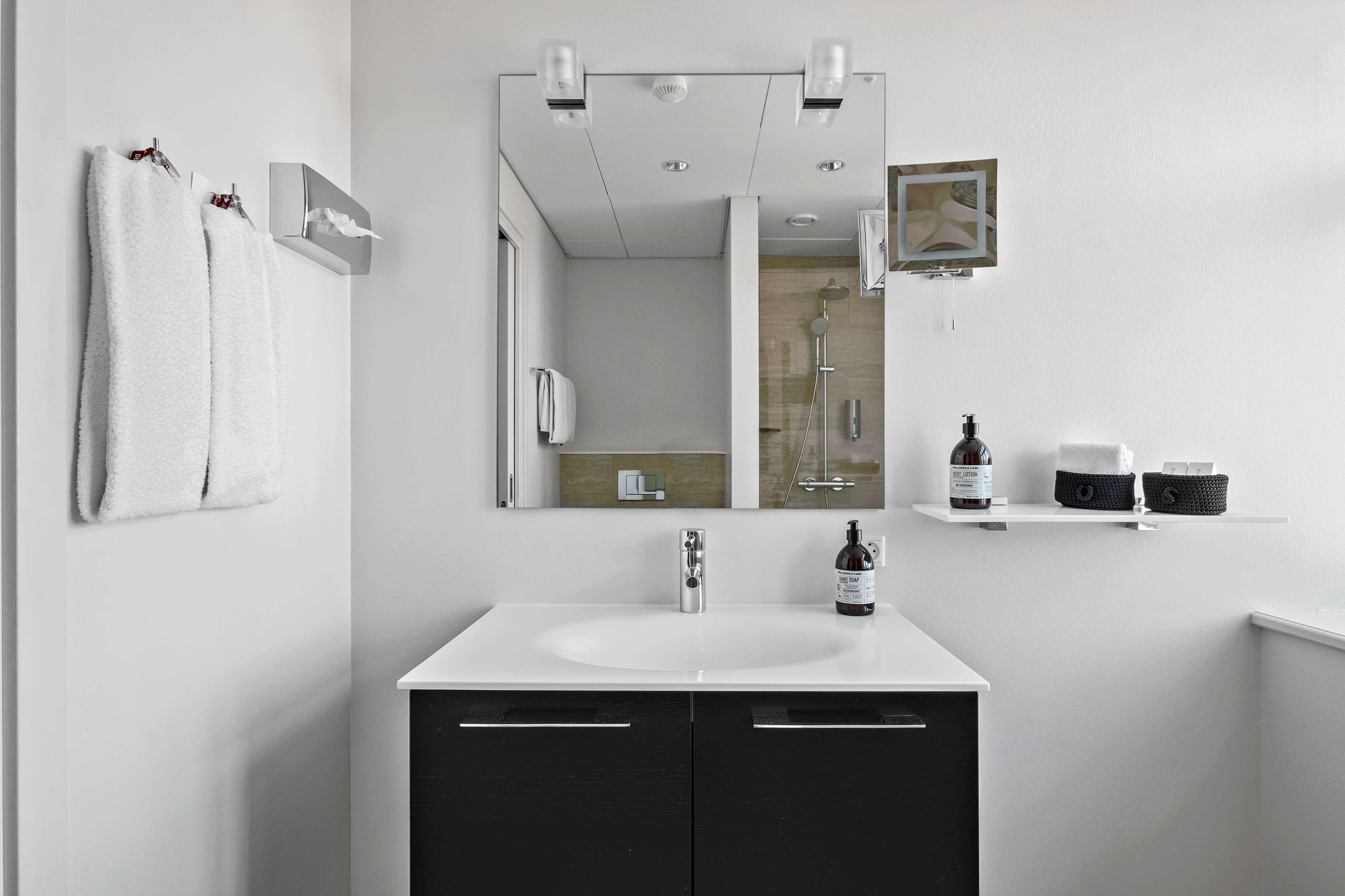 Un baño moderno con lavamanos blanco, muebles bajos negros, un espejo grande y un perchero para toallas.