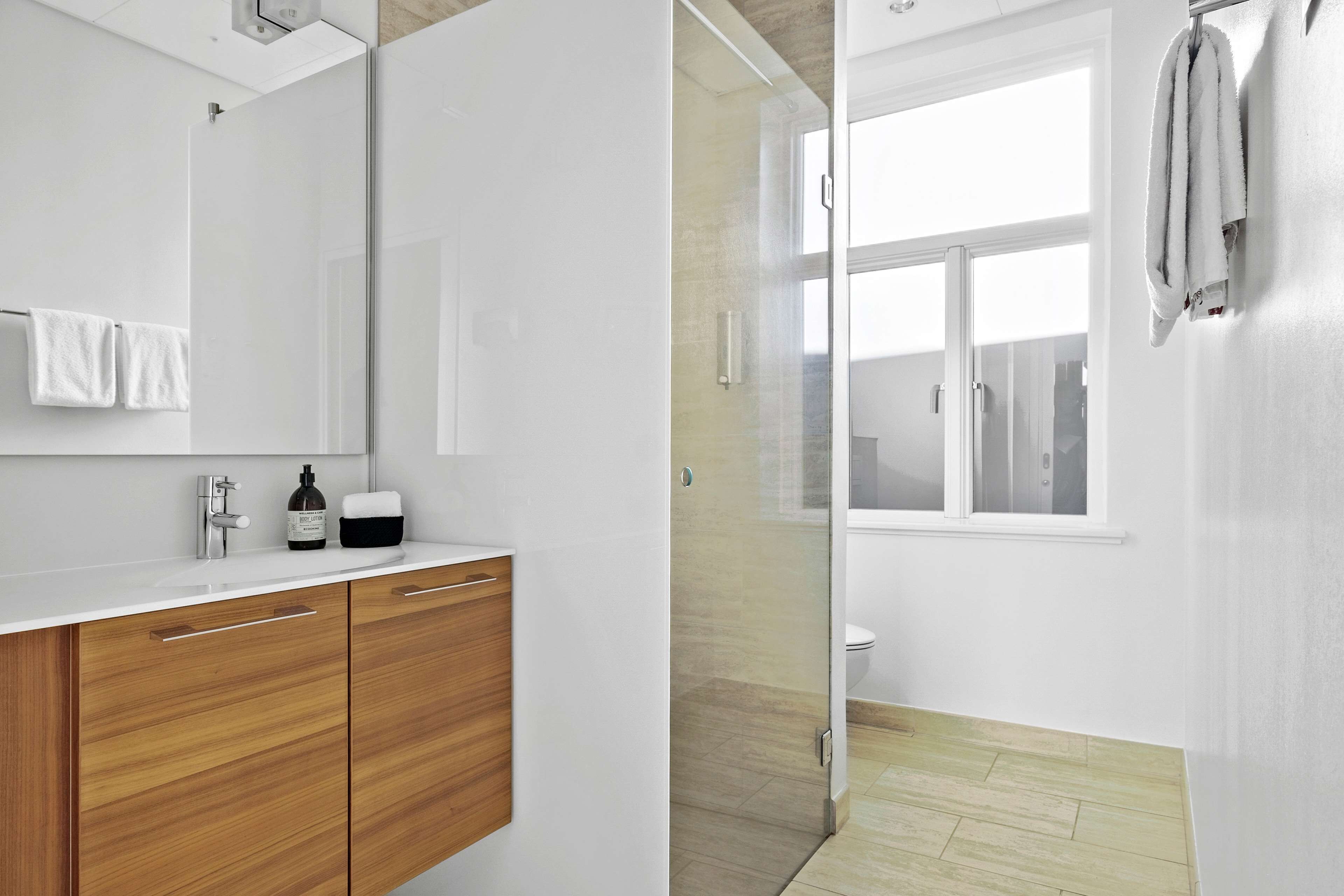 A modern bathroom with a glass shower enclosure, a white sink with wooden cabinets, and a window providing natural light.