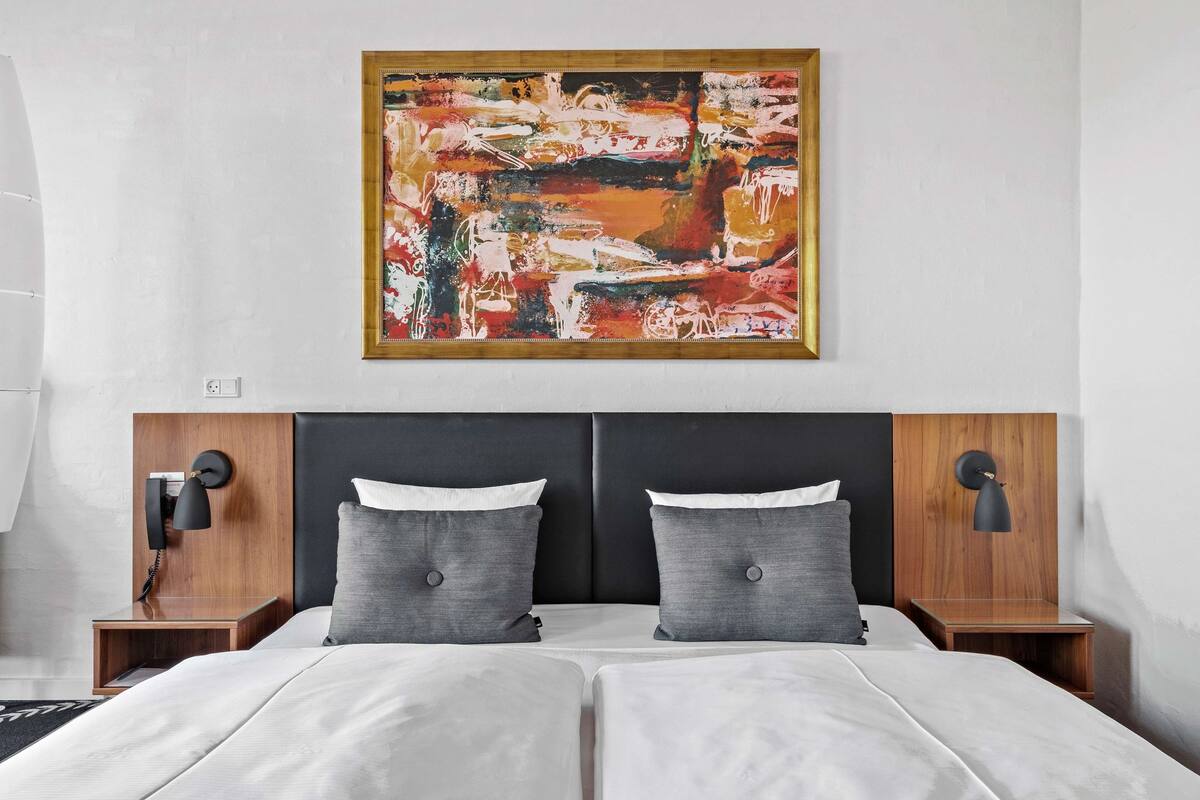 Una habitación de hotel moderna con un cuadro abstracto grande, una cama doble con ropa de cama gris, dos mesitas de noche con lámparas y un cabecero de madera.