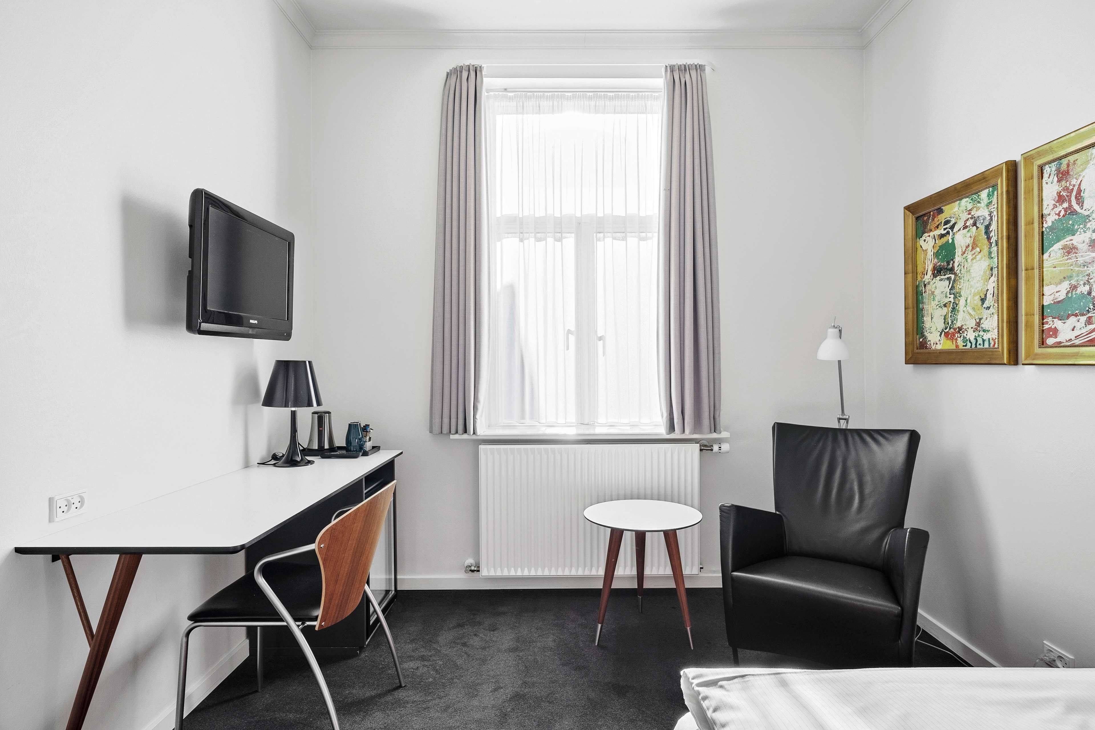A hotel room with a desk, chair, bed, TV, window with curtains, radiator, and artwork on the wall.