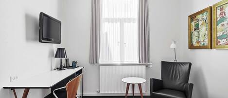 A hotel room with a desk, chair, bed, TV, window with curtains, radiator, and artwork on the wall.