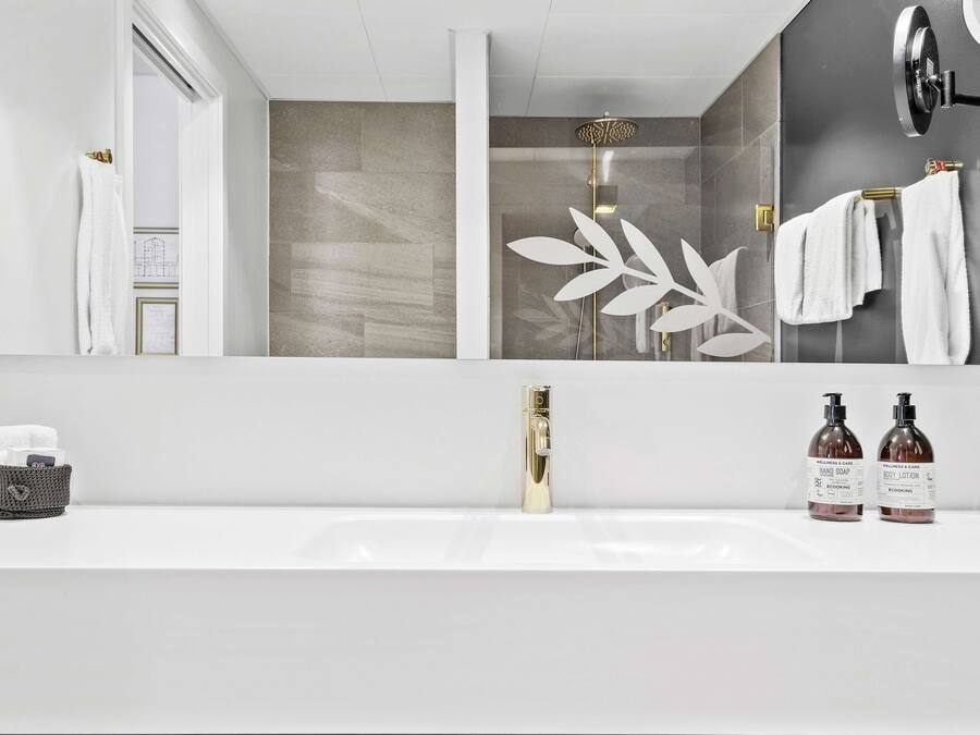 A modern bathroom with a large mirror, a shower with a gold faucet, and two bottles of body lotion on the sink.