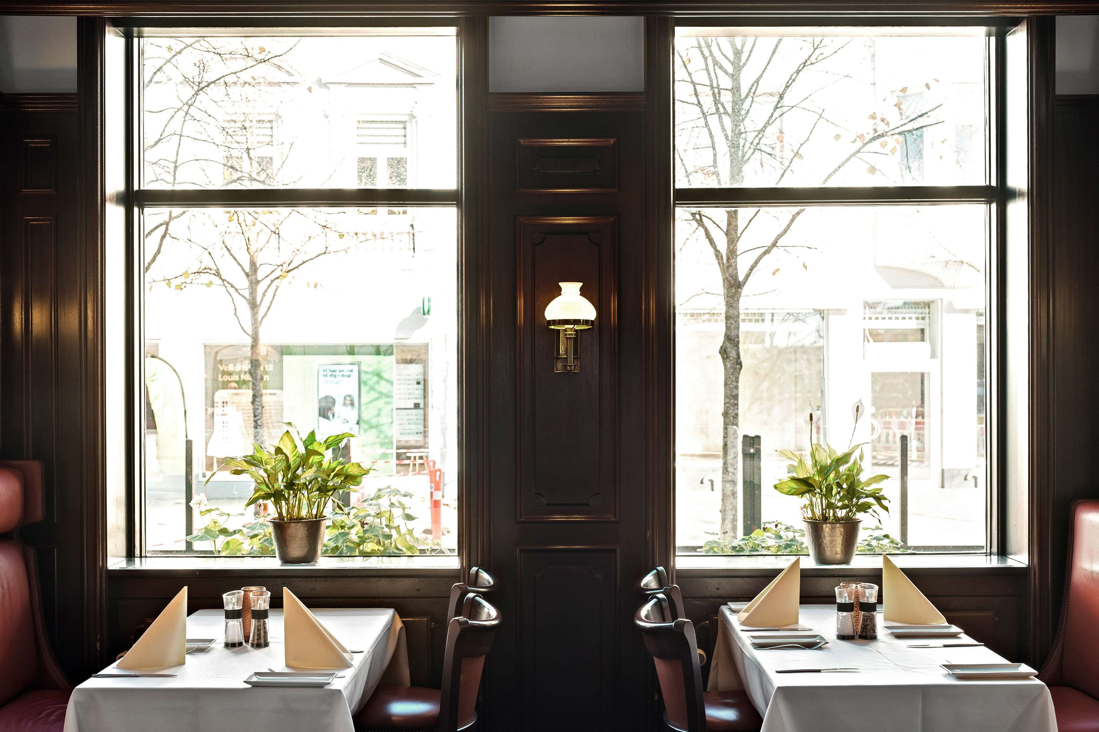 Un restaurante con una larga mesa dispuesta para dos, ventanas amplias con plantas en macetas y una lámpara fijada a la pared.