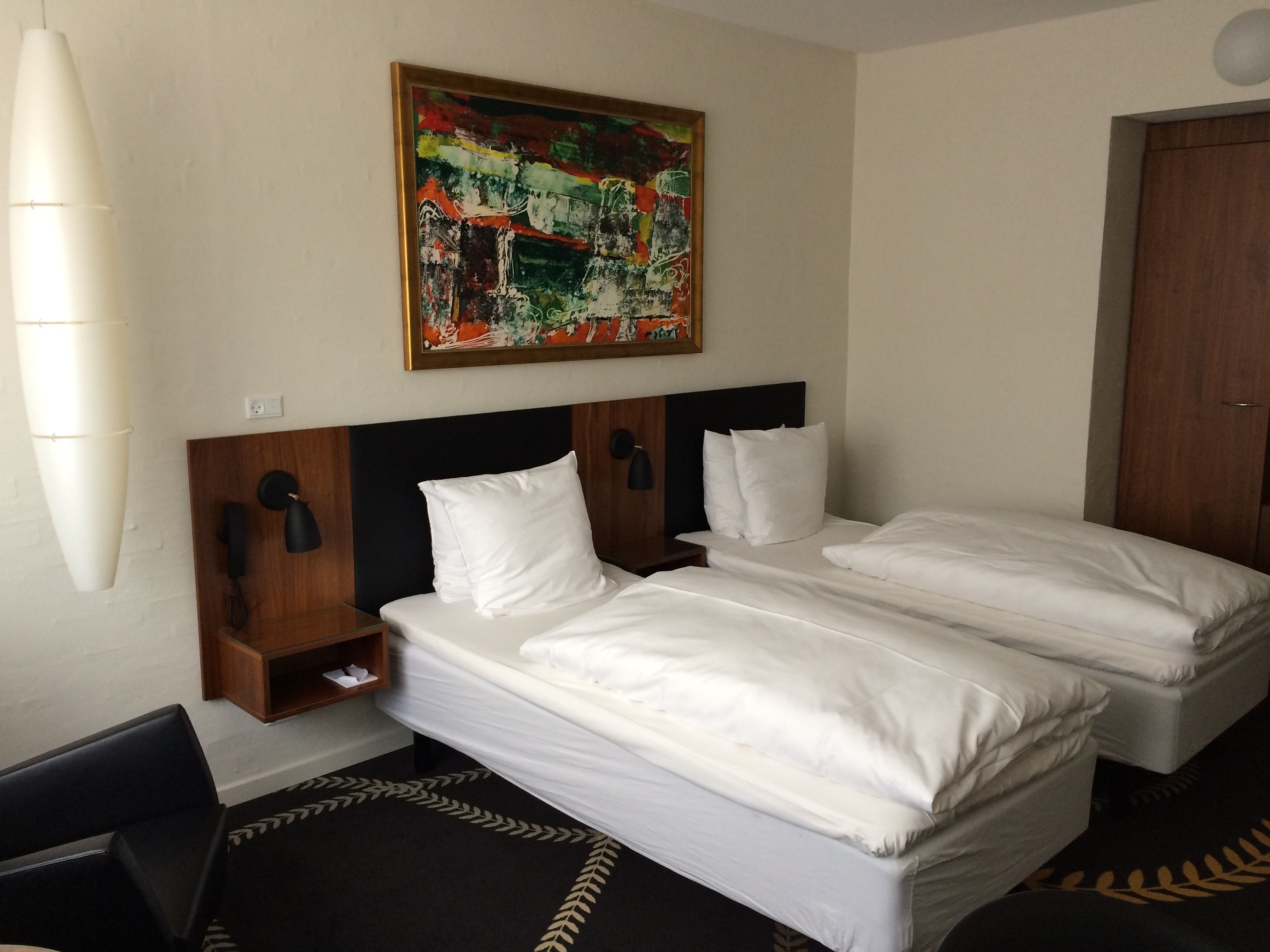 A hotel room with two beds, a painting on the wall, a bedside table with a lamp, and a door.